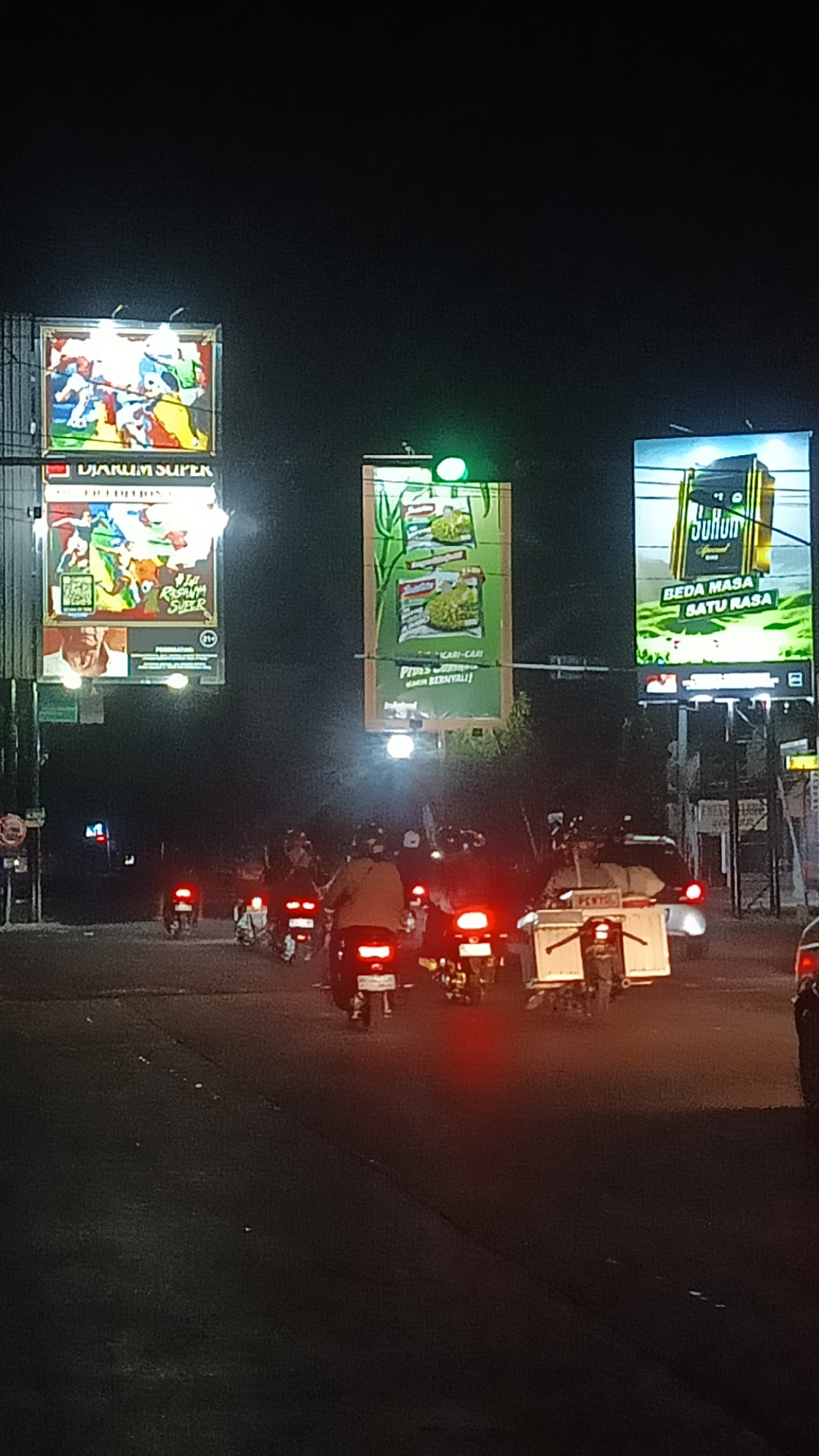 A night scene captured in Ngrampal, Indonesia, shows a busy road with several motorcycles and a sidecar motorcycle navigating the street. The environment is illuminated by various billboards advertising consumer products. On the left, a billboard displays "DJARUM SUPER" with colorful graphics of people in action. To the right, another billboard features images of Indomie instant noodles with the text "KARI-CARI" and "PIKIR KARI-CARI PASTI BERNILAI!". Further to the right, a billboard for "Suryam" cigarettes is visible, with the tagline "BEDA MASA SATU RASA". The road appears to be a main thoroughfare, with traffic light signals also present, suggesting a potentially active intersection. The overall atmosphere is one of urban nighttime activity.