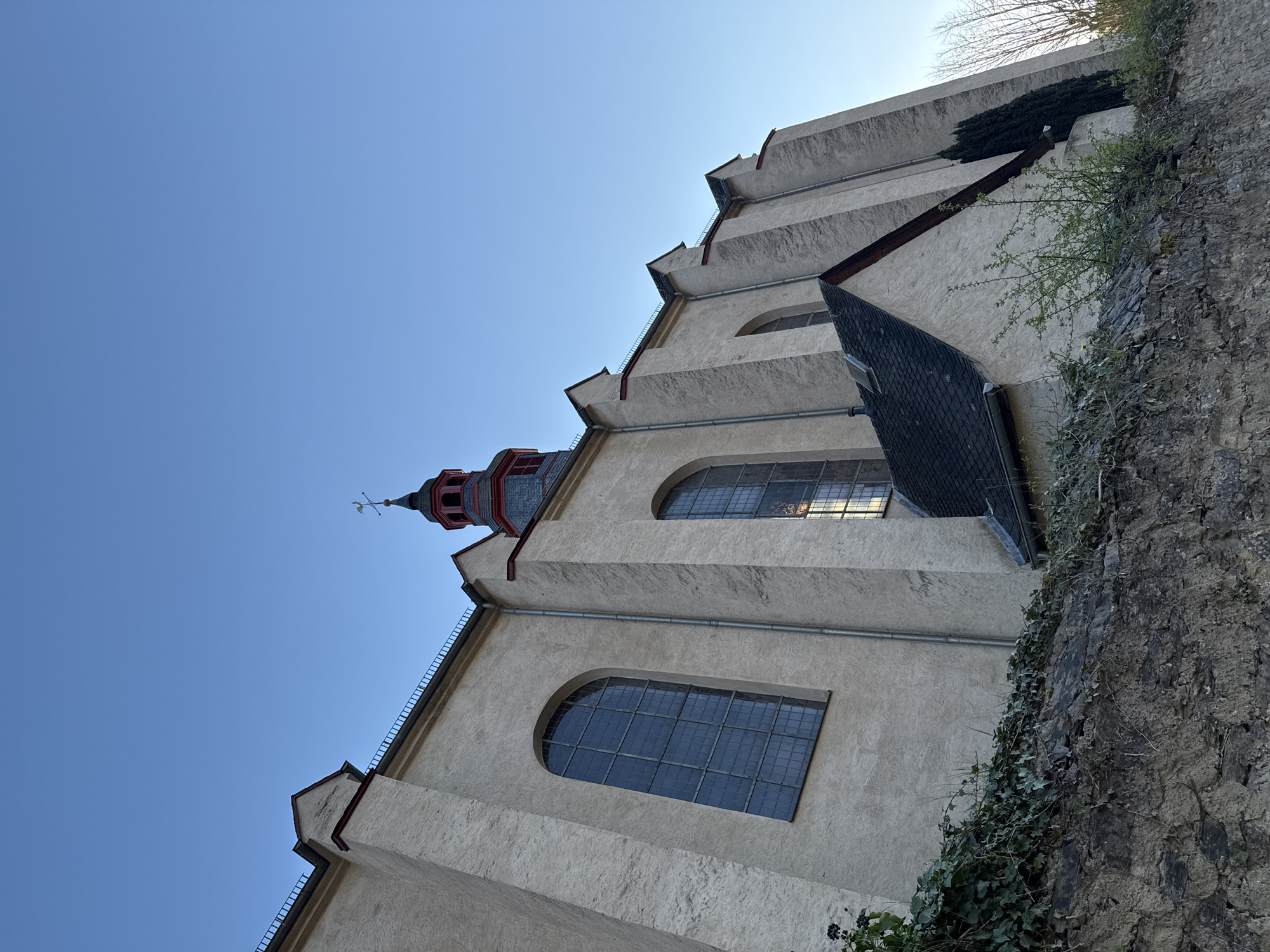 The image depicts a low-angle view of the Church of St. Joseph in Beilstein, Germany. The church's facade is visible, including its stepped exterior design with large arched windows and a spire topped with a weather vane. A stone wall covered with vegetation flanks the right side of the church. The sky is clear and blue. A section of a tiled roof extends from the church structure.