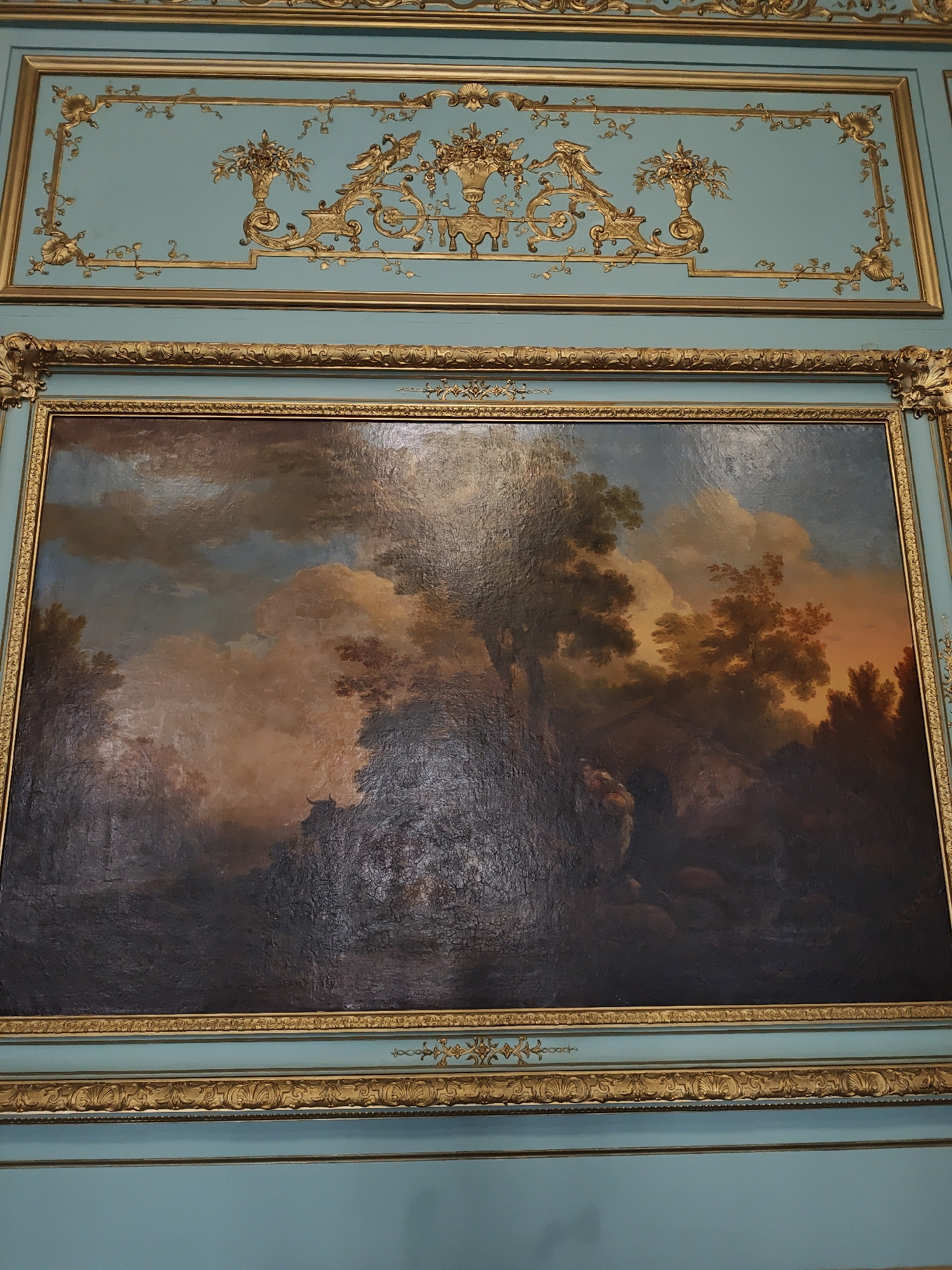 The image depicts a gilded wooden decorative panel above a framed painting. The panel features intricate gold ornamentation against a light teal background, with repeating floral motifs and a central cartouche containing what appears to be a deer and birds. Below this, a large, rectangular painting is framed by an ornate gold border. The painting itself is a landscape scene with dramatic, cloudy skies transitioning to warm hues of orange and yellow near the horizon. Dark, shadowy trees and foliage dominate the foreground, with hints of what could be figures or animals within the dense shadows. The painting is displayed on a light teal wall, with further gold decorative trim below the painting's frame. This scene is located in the Potocki Palace in Lviv, Ukraine.