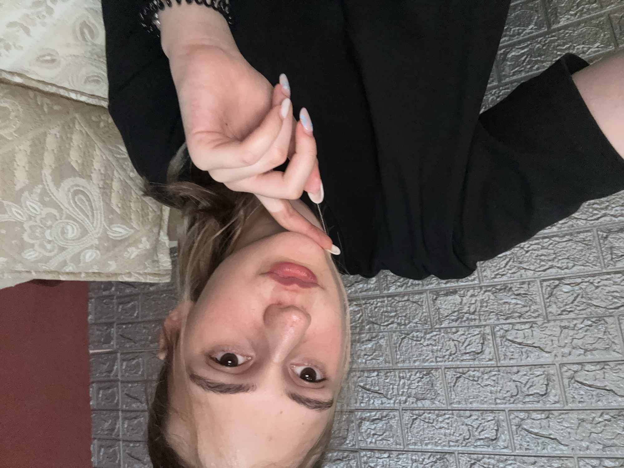 A young woman is photographed from a slightly upward angle. Her head is tilted downward towards the bottom of the frame. She is Caucasian with light skin, brown eyes, and fair hair partially pulled back. She is wearing a black top or dress. Her left hand is raised, with her fingers touching her chin. She wears a beaded bracelet on her left wrist and has manicured nails with a white and light blue design. The background behind her head features a gray brick-like wall. To the left, there is a floral patterned cushion. A red object is partially visible near the cushion. There is no evidence suggesting the location is Panagyurishte, Bulgaria.