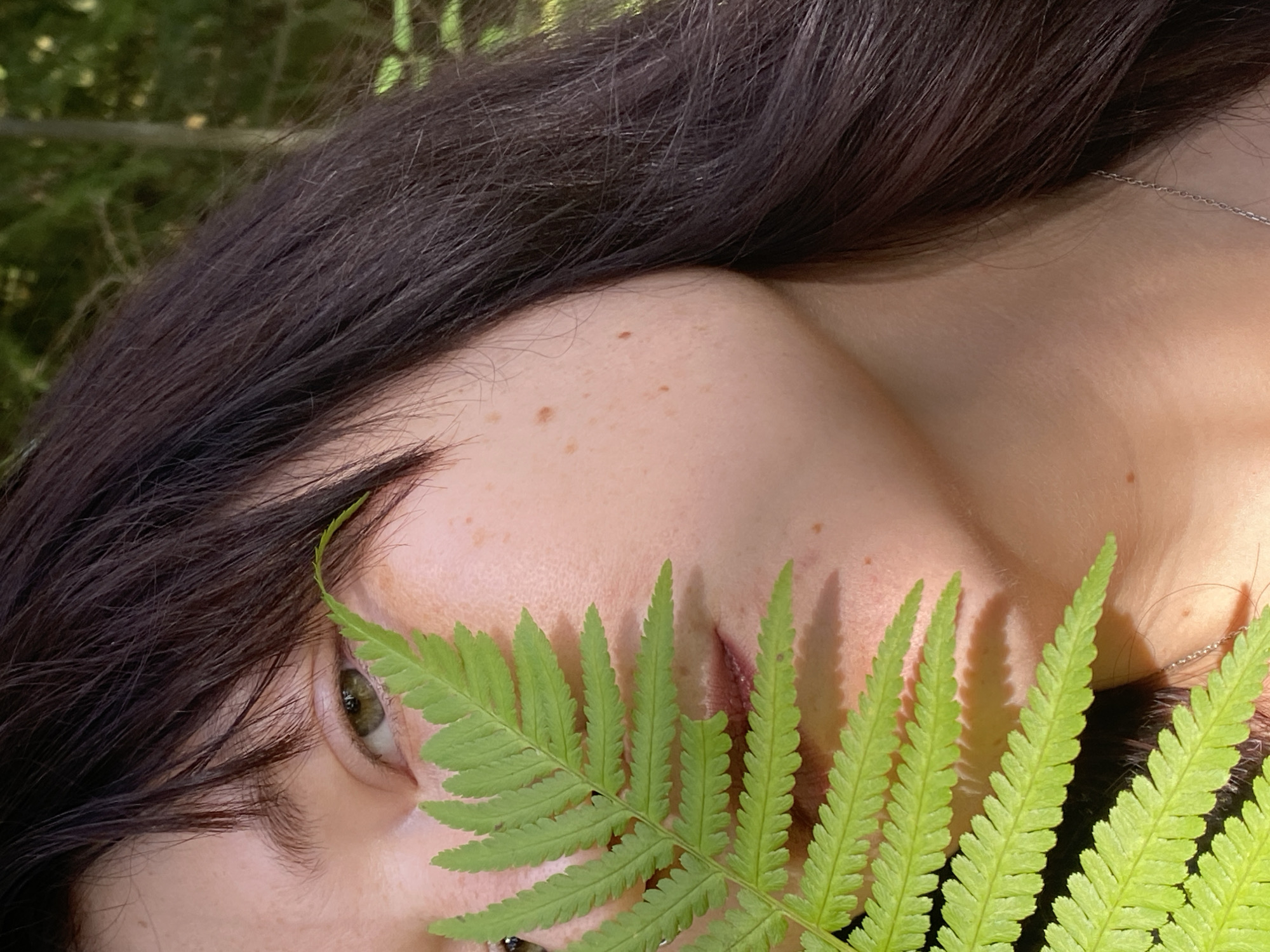 A close-up, eye-level shot shows a person's face from the nose up, partially obscured by a large, vibrant green fern frond. The subject has fair skin with freckles, dark brown hair, and a visible portion of their green eye. The background consists of out-of-focus greenery, suggesting an outdoor natural setting, possibly a forest or park. The lighting appears to be natural sunlight, casting shadows from the fern onto the subject's face. There is no identifiable location in the city of Фоки, Russia.