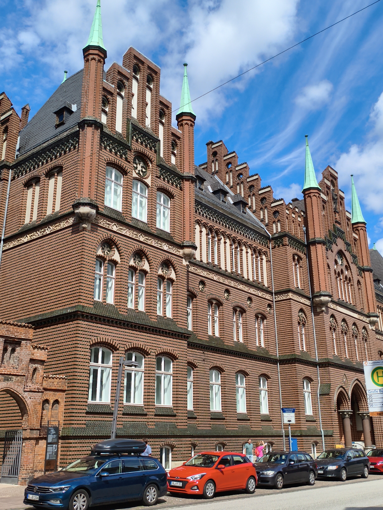 This is a full-body, slightly low-angle, eye-level shot of a long, ornate, red-brick building in Lübeck, Germany. The building features multiple pointed green spires, arched windows with white frames, and intricate decorative brickwork. In the foreground, several cars are parked along the street. On the left, a blue station wagon with a roof box is parked. To its right, an orange hatchback is parked, followed by two dark gray station wagons, and another red vehicle is partially visible on the far right. A traffic sign with a green "H" symbol and German text is visible on the right side of the image. The sky is blue with scattered white clouds. A few people are visible walking on the sidewalk in front of the building and near the parked cars. The overall scene appears to be a street in an urban environment on a sunny day.