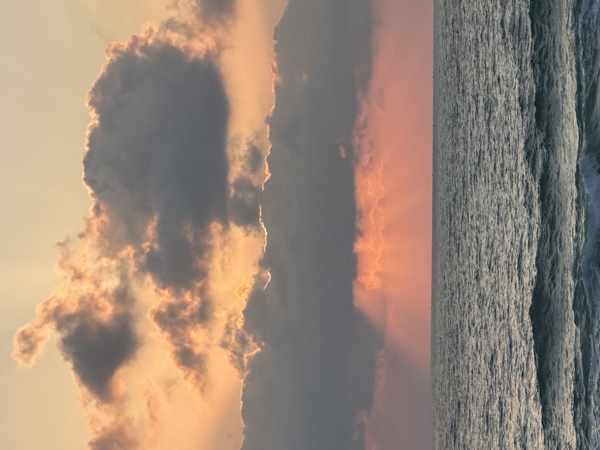 The image depicts a serene seascape at sunset, likely on the coast of Mate Asher Regional Council, Israel. The sky is a canvas of warm hues, dominated by soft oranges and pinks, with dramatic clouds casting shadows and allowing golden rays of sunlight to pierce through. The sea is a deep, textured blue, with gentle waves rolling towards the shore. The horizon is a clear, thin line separating the sky from the water, with faint silhouettes of what appear to be boats or buoys scattered across the surface. There are no people or specific structures visible in the frame, emphasizing the natural beauty of the coastal environment.