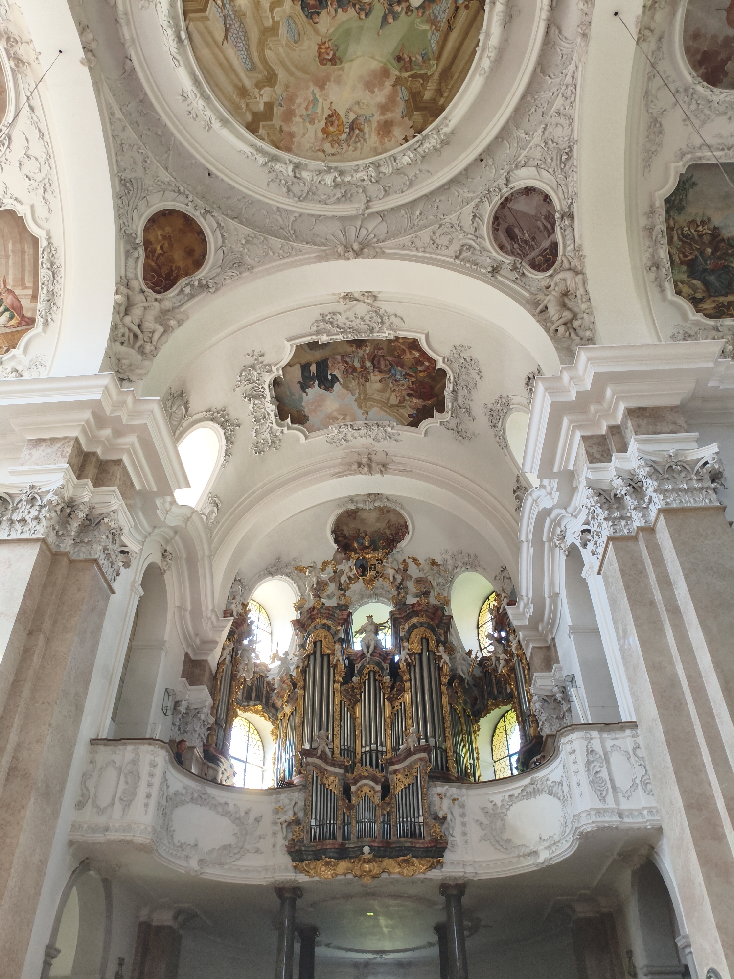 The image captures the opulent interior of a church, likely the St. Mang's Abbey in Füssen, Germany, due to its distinctive Baroque architecture. The ceiling is elaborately decorated with frescoes depicting biblical scenes and angelic figures, surrounded by ornate white stucco work. Large pillars with marble-like detailing flank the central nave. Dominating the mid-ground is a magnificent pipe organ, a golden and dark wood structure adorned with sculptures and intricate carvings. The organ is situated on a balcony that extends from the wall, featuring detailed white stucco railings and cherubic figures. Behind the organ, arched windows with stained glass allow light to filter into the sanctuary. A single person can be seen in the organ loft, appearing to be operating the instrument. The overall impression is one of grandeur and artistic mastery, characteristic of Baroque religious architecture.