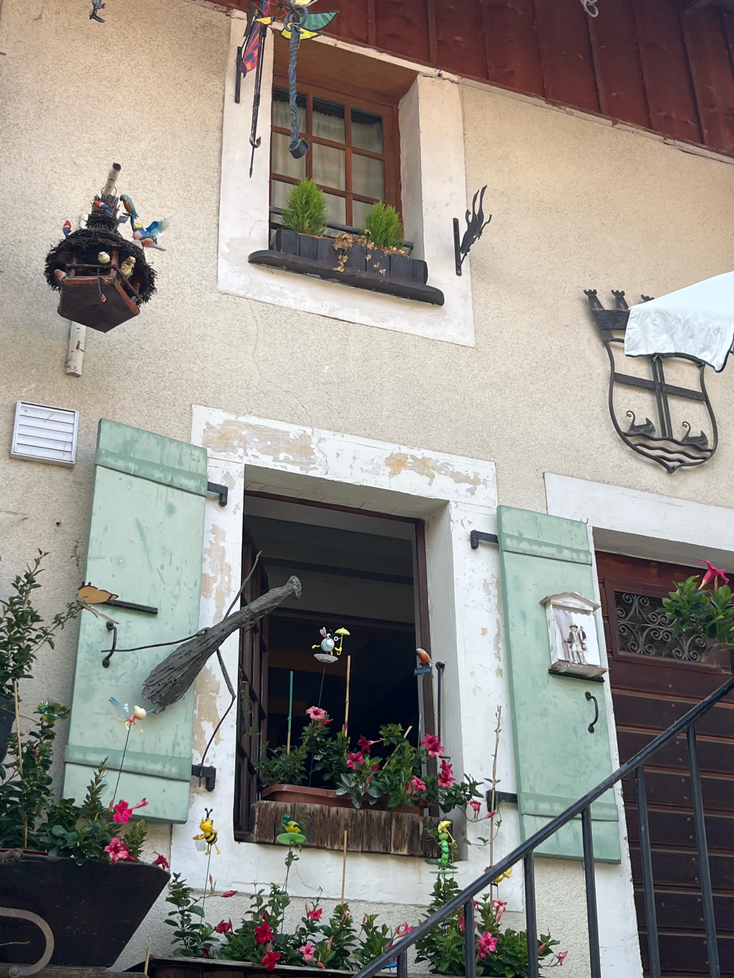 The image shows the exterior of a building in the village of Yvoire, France, with decorative elements and plants. The building has a light-colored stucco exterior and a dark wooden roofline.
In the upper left, a birdhouse with several small bird figures is hanging from a pole. The birdhouse itself is dark brown with a thatched roof and is adorned with various colorful bird figurines. Above this, a decorative mobile with colorful panels and a black cord hangs from the building.
To the right of the birdhouse, a window with a wooden frame and multi-paned glass is visible. A dark wooden planter box sits below the window, containing two small green plants and some dried leaves.
A large, open window with light green shutters is prominent in the lower half of the image. The shutters are partially open, revealing a glimpse of the interior. A large, gnarled wooden branch is positioned across the lower part of this open window. Hanging from sticks in front of the window are several garden ornaments, including a blue dragonfly and a yellow bee. A small red bird figurine is perched on the window frame to the left.
To the right of the open window, a closed window with light green shutters is visible. On the shutter, a small decorative diorama depicting two figures in traditional attire is mounted within a white architectural frame. Further to the right, a dark wooden door with ornate metal grilles is present. Above the door, a black metal coat of arms with a shield shape and boat-like figures is mounted on the wall. A white tablecloth covers part of the coat of arms.
In the foreground, a black metal railing runs diagonally across the lower right portion of the image, suggesting a staircase. Pots of pink flowering plants are arranged in front of the building and on a lower level. A small green caterpillar-like garden ornament is visible amongst the plants. A white vent is mounted on the stucco wall to the left of the open window.
The overall scene suggests a charming, decorated facade in a European village setting.
