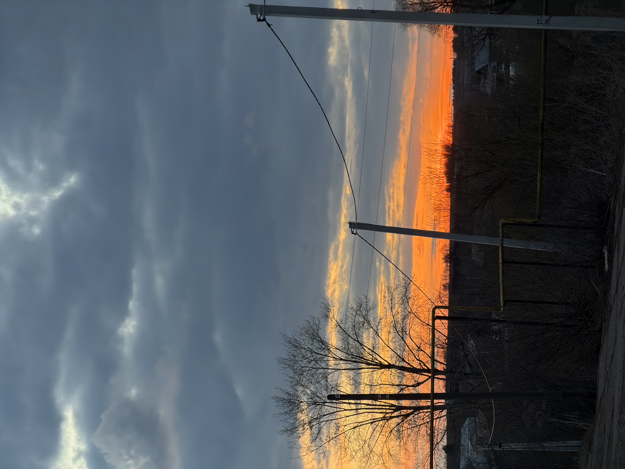 This image captures a landscape scene in Кварса, Russia, at either sunrise or sunset. The sky is a dominant feature, filled with extensive dark grey clouds that cover much of the upper portion of the frame. Below these heavy clouds, a vibrant band of orange and yellow light illuminates the horizon, indicating the position of the low sun.
In the foreground and midground, numerous bare-branched trees are silhouetted against this bright horizon, their intricate network of branches clearly visible. Utility poles with power lines crisscross the frame, adding a man-made element to the natural setting. Several yellow gas pipes are also visible, running horizontally across different parts of the scene. The ground appears to be an unpaved road or dirt path.
No people or specific activities are present, giving the scene a quiet and still atmosphere. The combination of the dramatic, cloud-filled sky, the intense orange light, and the silhouetted bare trees suggests the chill of late autumn or winter. There is no visible text in the image.