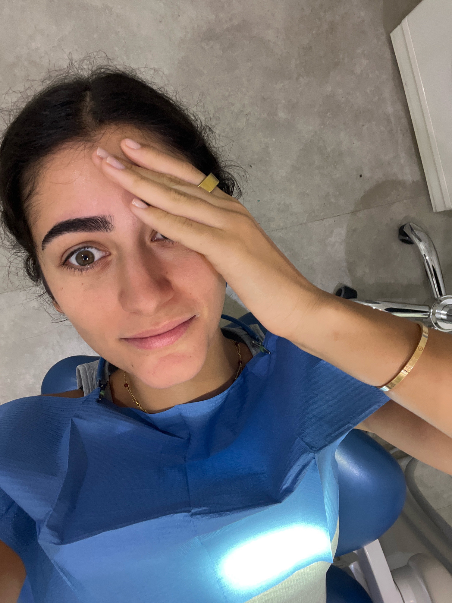 A young woman with dark hair and brown eyes is seated in what appears to be a dental chair, evidenced by the blue dental bib she is wearing and the dental chair itself, partially visible. Her right hand is covering her left eye, while her right eye is open and looking towards the camera. She is wearing a gold ring on her right index finger and a gold bracelet on her right wrist. The background consists of a gray, textured wall and a white object, possibly a cabinet or counter, on the right. The lighting suggests an indoor environment. The image was captured in Porto, Portugal.
