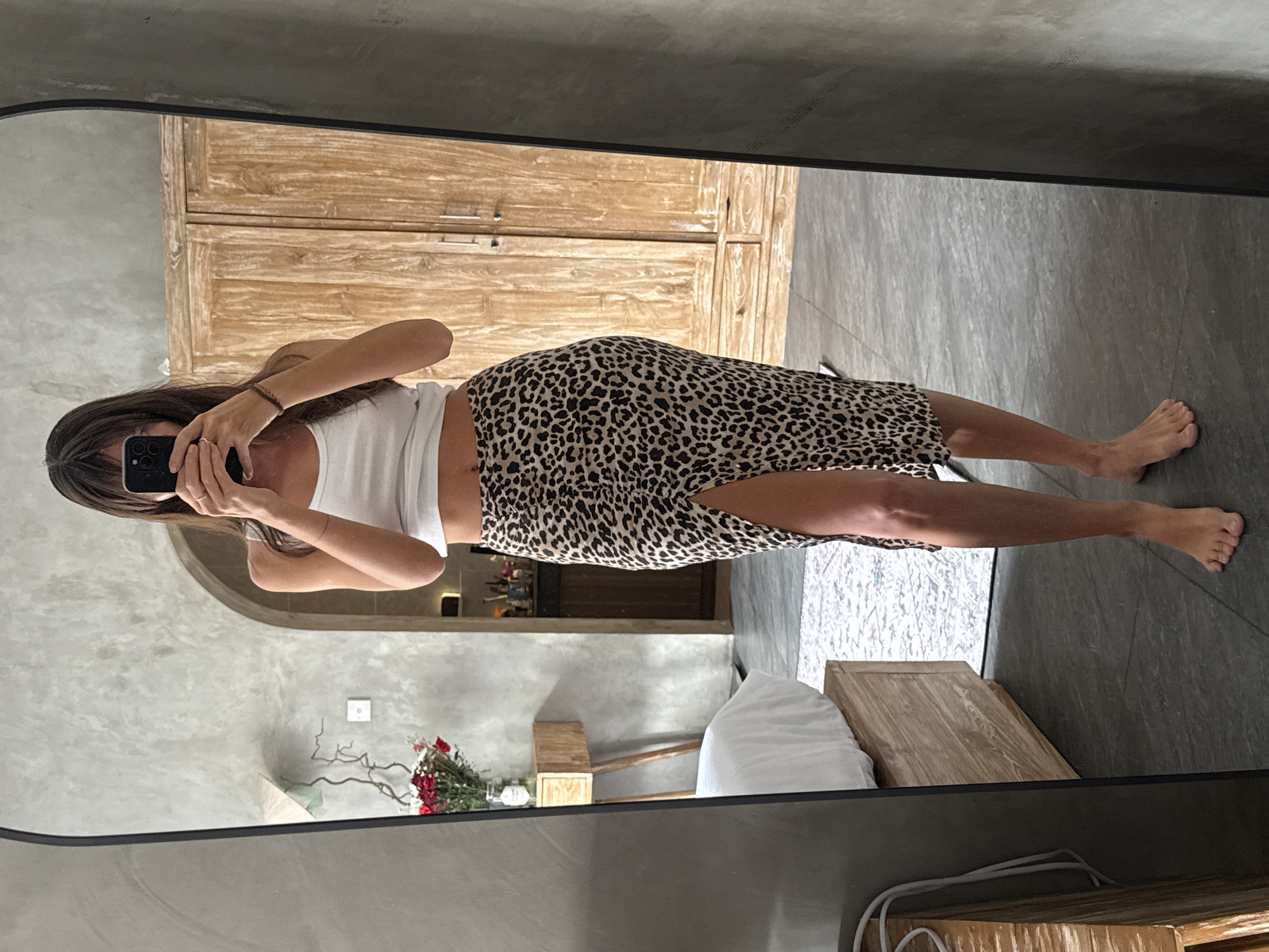 A woman is seen in a mirror taking a selfie in what appears to be a room in North Kuta, Indonesia. The woman is wearing a white tank top and a leopard print midi skirt with a slit. Behind her in the mirror's reflection is a wooden cabinet and part of a bed with white linens. A bouquet of red flowers is also visible in the reflection, along with walls finished with a concrete-like texture. The floor is tiled in a dark color.