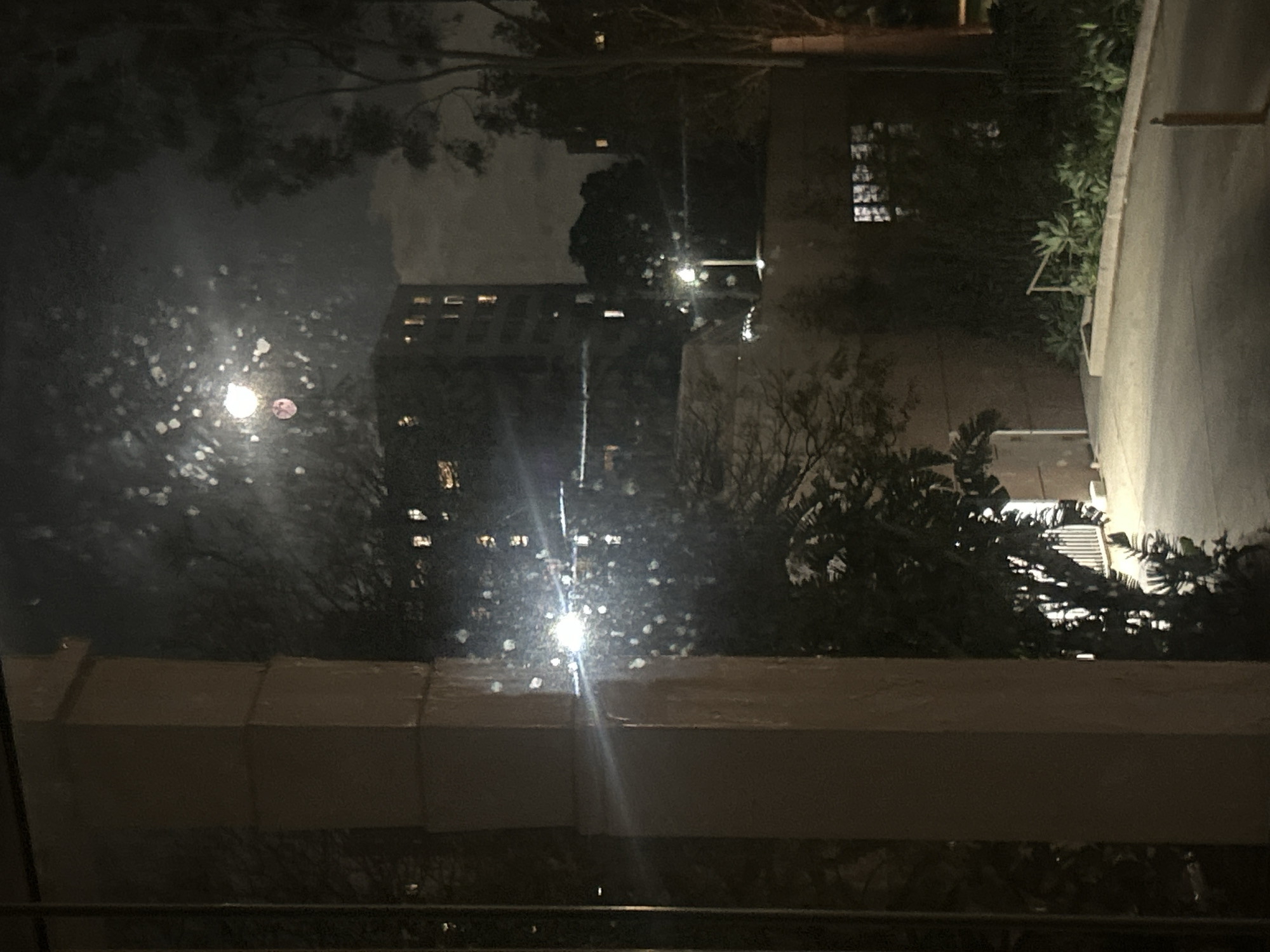 A low-angle night shot shows a rain-streaked window looking out at a cityscape. The window is framed by a white or light-colored concrete structure, possibly a balcony or ledge. Behind the window, buildings with lit windows are visible. Streetlights are also visible and blurry due to the rain on the window. The overall scene is dark and somewhat obscured by the rain. The location is likely in a residential area of Los Angeles, based on the dense urban setting and building architecture.