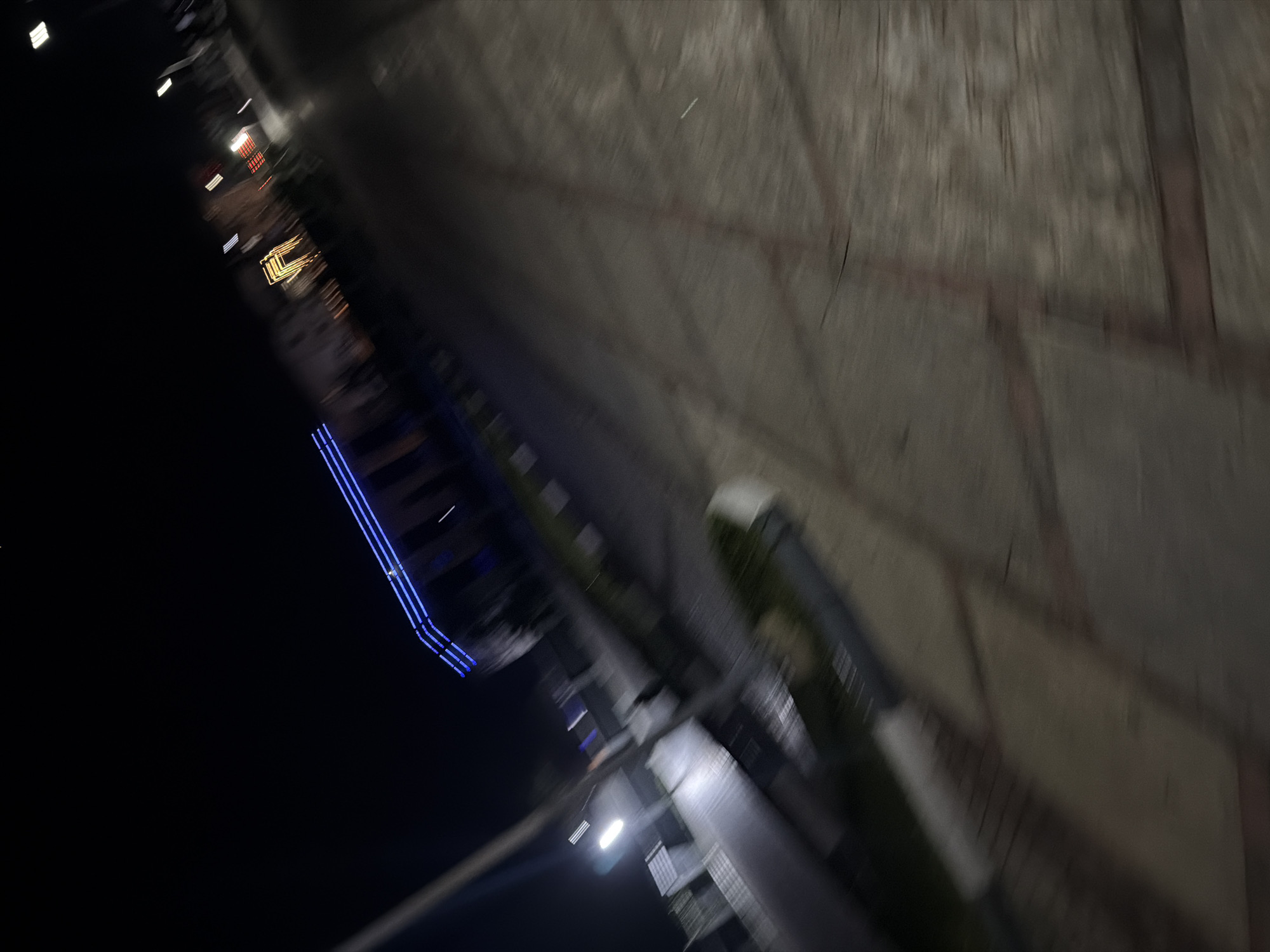The image, taken at night in Bukuru, Nigeria, is heavily motion blurred. The scene appears to be an outdoor public space, possibly a walkway or plaza. In the background, there are lights from buildings, including what looks like blue neon lights outlining a structure and yellow lights from a doorway or window. Streetlights are also visible, emitting a white glow. The foreground and middle ground are dominated by a blurred paved surface, with faint lines suggesting patterns or divisions in the pavement. There are also blurred shapes that could be benches or planters. The overall impression is a dark, urban environment at night, with artificial lighting creating streaks and blurs due to the motion of the camera.