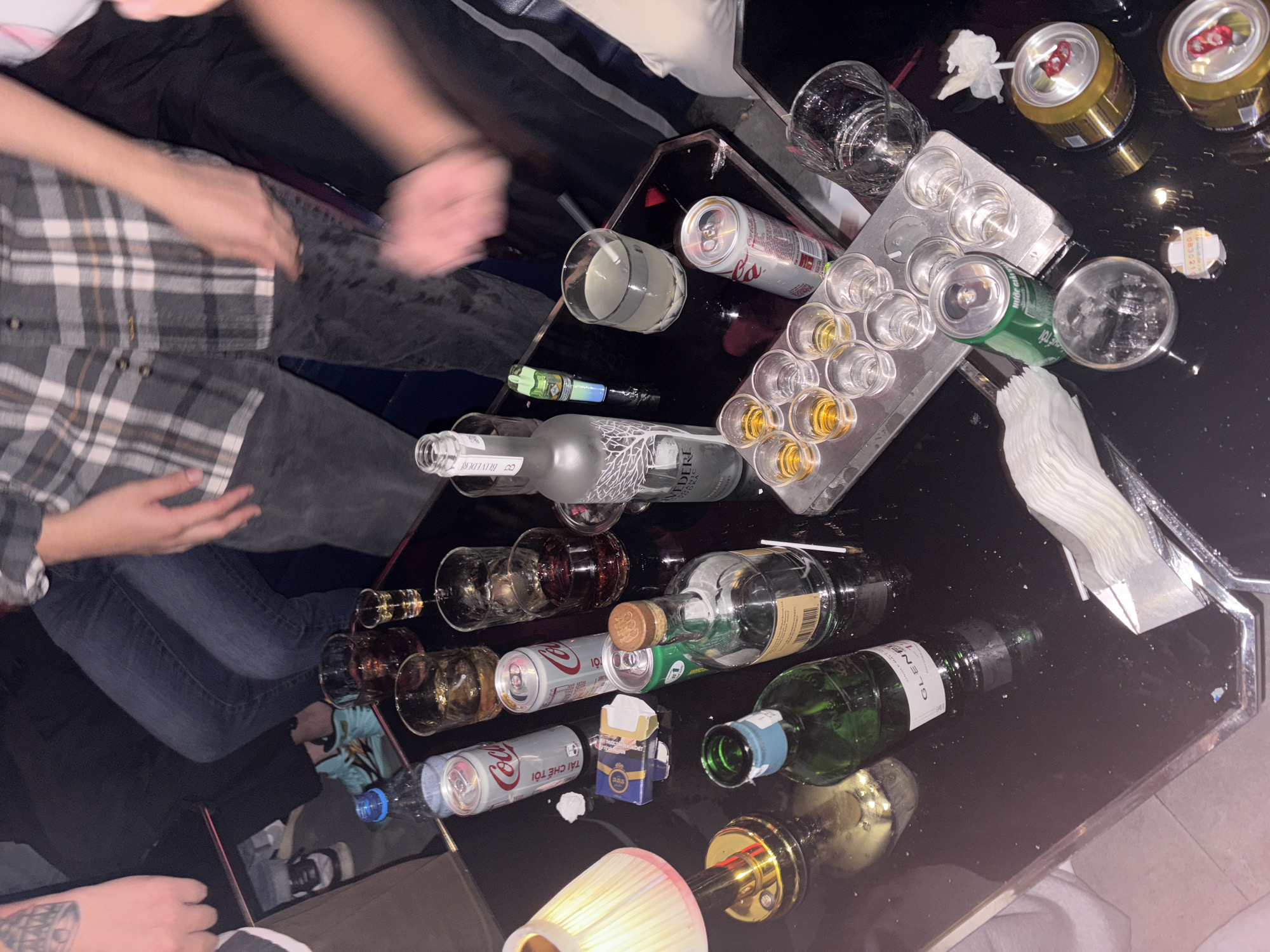 The image depicts a dimly lit, indoor scene, likely a bar or private gathering, with a focus on a table laden with alcoholic beverages and associated items. A group of individuals, partially visible, are present, their legs and arms blurred due to motion. The table surface is dark and reflective, covered with various objects including:
**Beverages:**
* Several cans of Coca-Cola.
* A bottle of Belvedere Vodka.
* A green bottle with a label partially obscured, appearing to be a spirit like Glenmorangie.
* Multiple shot glasses, some filled with amber liquid, others empty.
* Several larger glasses, one containing a clear beverage with a straw.
* Two open cans of a golden beverage with red pull tabs, possibly beer.
* A bottle of water.
**Other Objects:**
* A small, ornate lamp with a white shade, providing localized light.
* A pack of cigarettes.
* A lighter.
* A tray holding shot glasses.
* A stack of white napkins or paper towels.
* Various small, scattered white debris, possibly from the cigarettes or food.
**Environment:**
The background suggests a casual, possibly crowded setting, with dark seating and flooring. The overall impression is one of a relaxed and informal event involving alcohol consumption. While the image doesn't explicitly identify the location in Thủ Đức, Vietnam, the presence of Vietnamese text on one of the Coca-Cola cans ("Tết đến rồi") suggests it is indeed in Vietnam. The time appears to be evening or night, given the lighting conditions.