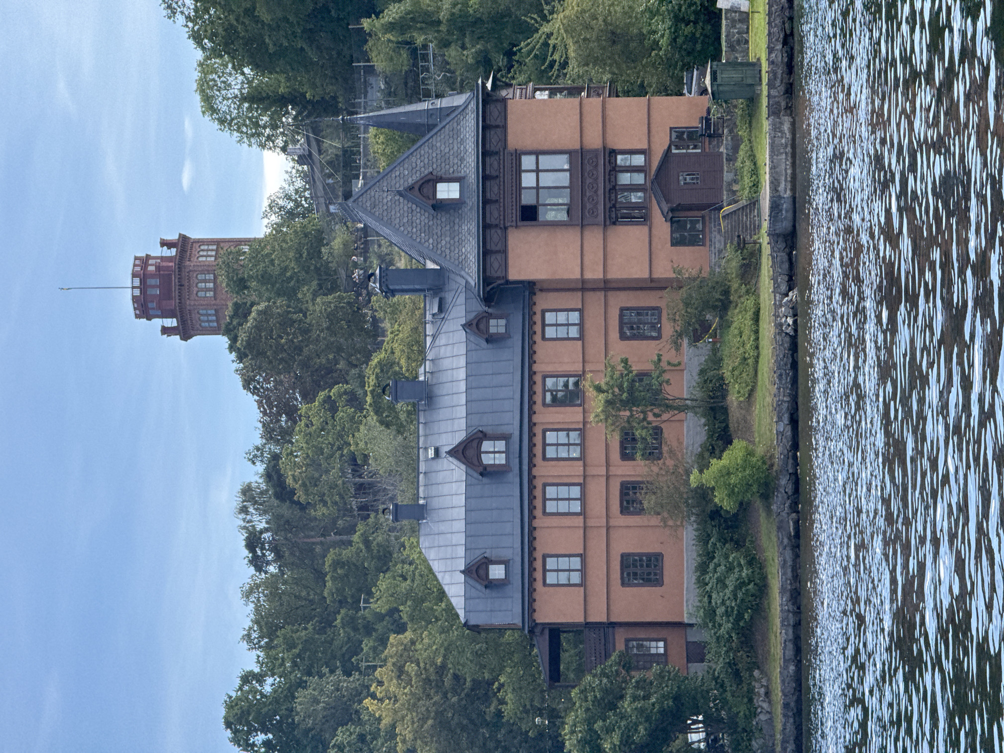This media file captures a view of a distinctive salmon-pink building with a steep grey roof, situated along the edge of a body of water. To the left, a tall, red brick tower with multiple levels and windowed sections stands prominently against a clear blue sky. The building has numerous windows, some with dark frames and mullions, and decorative wooden trim along its upper sections. The scene appears to be set in a somewhat natural environment with lush green trees bordering the building and the water's edge. The water itself is rippled, reflecting the light and giving a sense of movement. The overall impression is of a tranquil, perhaps historical, setting. No people are clearly visible. The time of day appears to be daytime, with the clear sky suggesting fair weather. The architecture of the building and the tower, coupled with the natural surroundings, hints at a European location, specifically Stockholm, Sweden, as indicated by the location context. There is no visible text in the image.