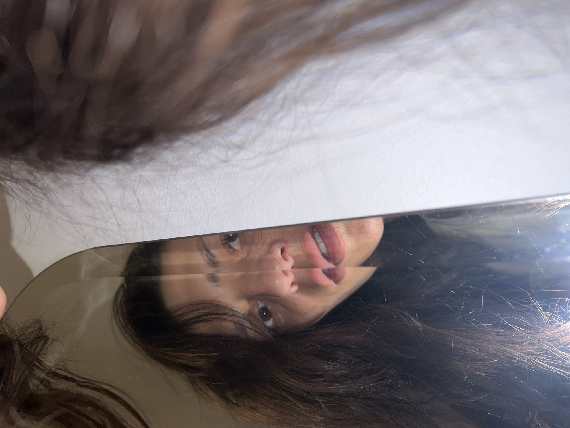 The image presents an unusual and fragmented perspective, bisected horizontally by a reflective surface, likely a mirror or a smartphone screen. The upper half of the image features a mass of dark brown hair, possibly belonging to the subject, partially obscuring a plain, light-colored background, which appears to be an indoor wall or ceiling.
Below the sharp, straight edge of the reflective surface, a person's reflection is prominently displayed. The reflection shows the face of a woman, appearing upside down from the viewer's perspective, framed by more of her dark brown, wavy hair that flows downwards in the reflection. Her facial features are captured in close-up: one dark eye is visible and looks directly towards the viewer, while the other is partially obscured by hair. Her nose and full lips are clearly discernible, with her mouth slightly open to reveal a hint of her teeth. Her expression is subtle and relaxed, perhaps a gentle pout. The lighting creates highlights on her hair and skin within the reflection.
The activity taking place is the woman observing her reflection, possibly while taking a selfie. The setting is indoors, implied by the plain background and diffused lighting. There are no specific time-of-day indicators or weather details. No distinct location cues are visible within the image itself. No text is visible.