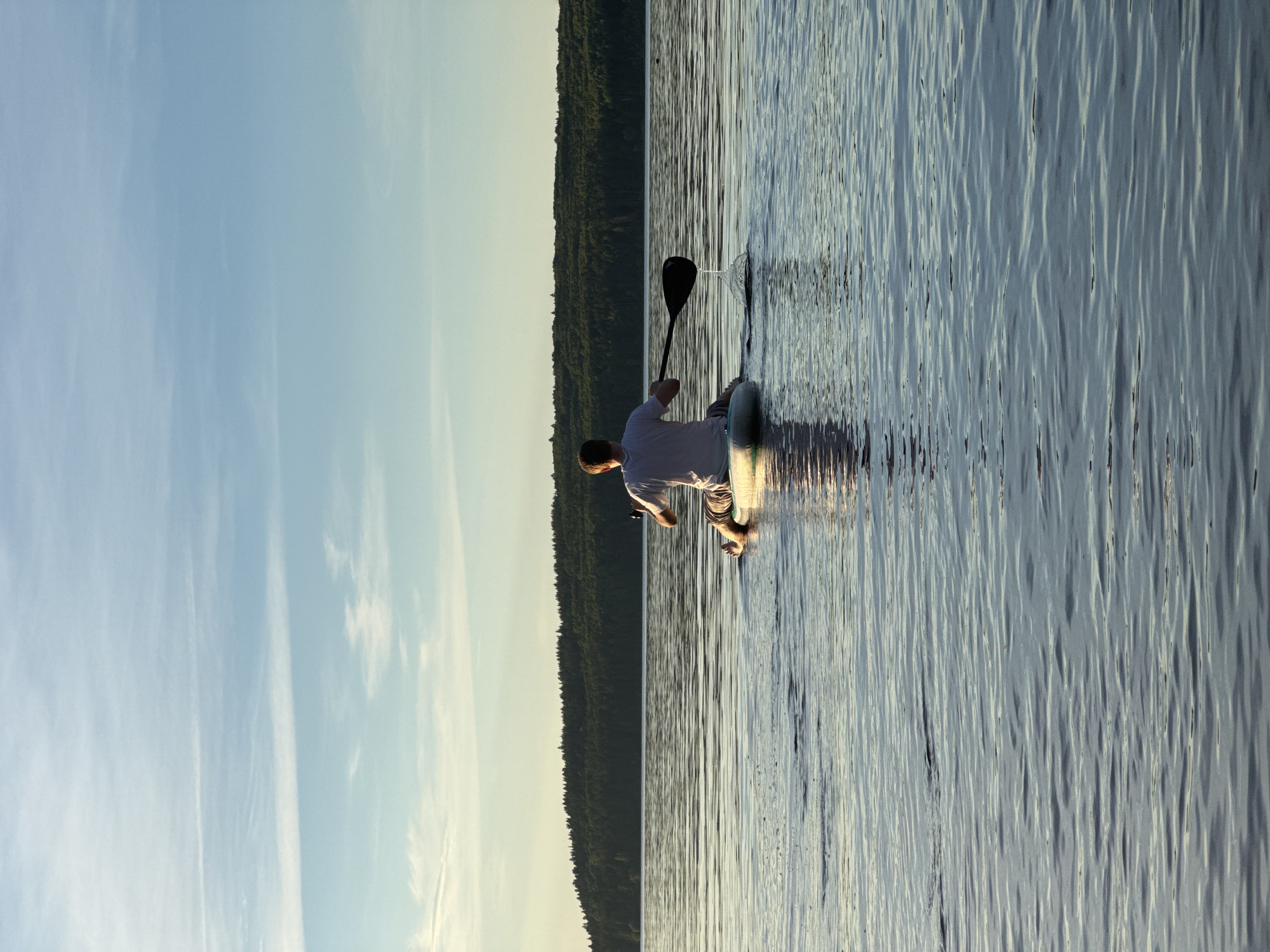 In this horizontal, eye-level shot, a single person is captured paddleboarding on a calm body of water. The individual, seen from behind, is wearing a white t-shirt and patterned shorts, their body angled as they prepare to paddle. Their stance suggests they are either about to begin their journey or are mid-stroke. The paddle, held firmly, dips into the water, creating a ripple that extends outwards.
The water itself is a prominent element, reflecting the sky and the distant shoreline with a rippled texture that gives it a shimmering appearance. The shoreline is densely covered with dark green trees, suggesting a natural, wooded environment. The sky above is a soft gradient of blue, transitioning to a pale yellow or white towards the horizon, with wispy white clouds scattered across it, indicating either early morning or late evening. The light is soft and diffused, contributing to a serene and peaceful atmosphere.
The scene evokes a sense of tranquility and solitude, with the vastness of the water and the quiet of the natural surroundings. The location context of Chaikovsky, Russia, places this serene moment in a specific geographical setting. No text is visible in the image. The time of day appears to be either sunrise or sunset, given the quality of the light and the sky's coloration.