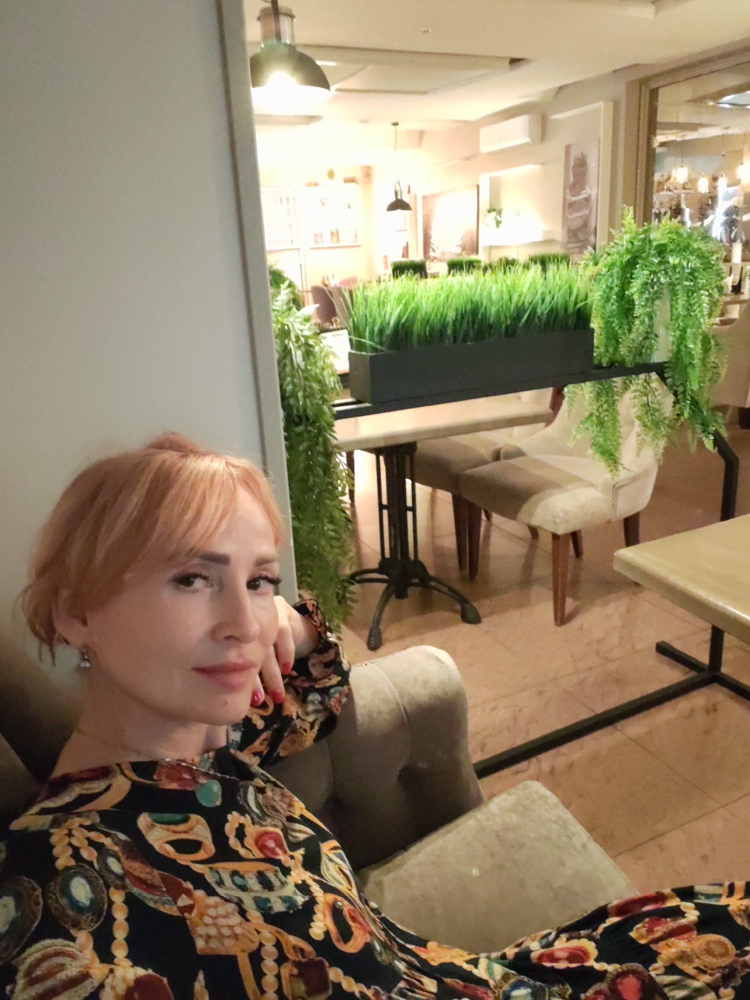 This media file is a selfie taken indoors. The main subject is a woman with short, reddish-blonde hair and fair skin. She is wearing a black garment with a colorful, ornate pattern featuring gold chains, jewels, and other decorative elements. She is looking towards the camera with a subtle smile and has her hand resting on her cheek. Her nails are painted red.
The setting appears to be a restaurant or cafe. In the background, there are tables and chairs. One chair is upholstered in a cream and taupe fabric. A long planter filled with lush green artificial grass is visible, with hanging artificial ferns on either side. Other decorative elements like framed artwork and lighting fixtures can be seen in the distance, suggesting an indoor environment that is lit artificially. The flooring is tiled in a warm, earthy tone. The overall atmosphere seems relaxed and casual. There is no visible text in the image. The time of day cannot be determined from the image.