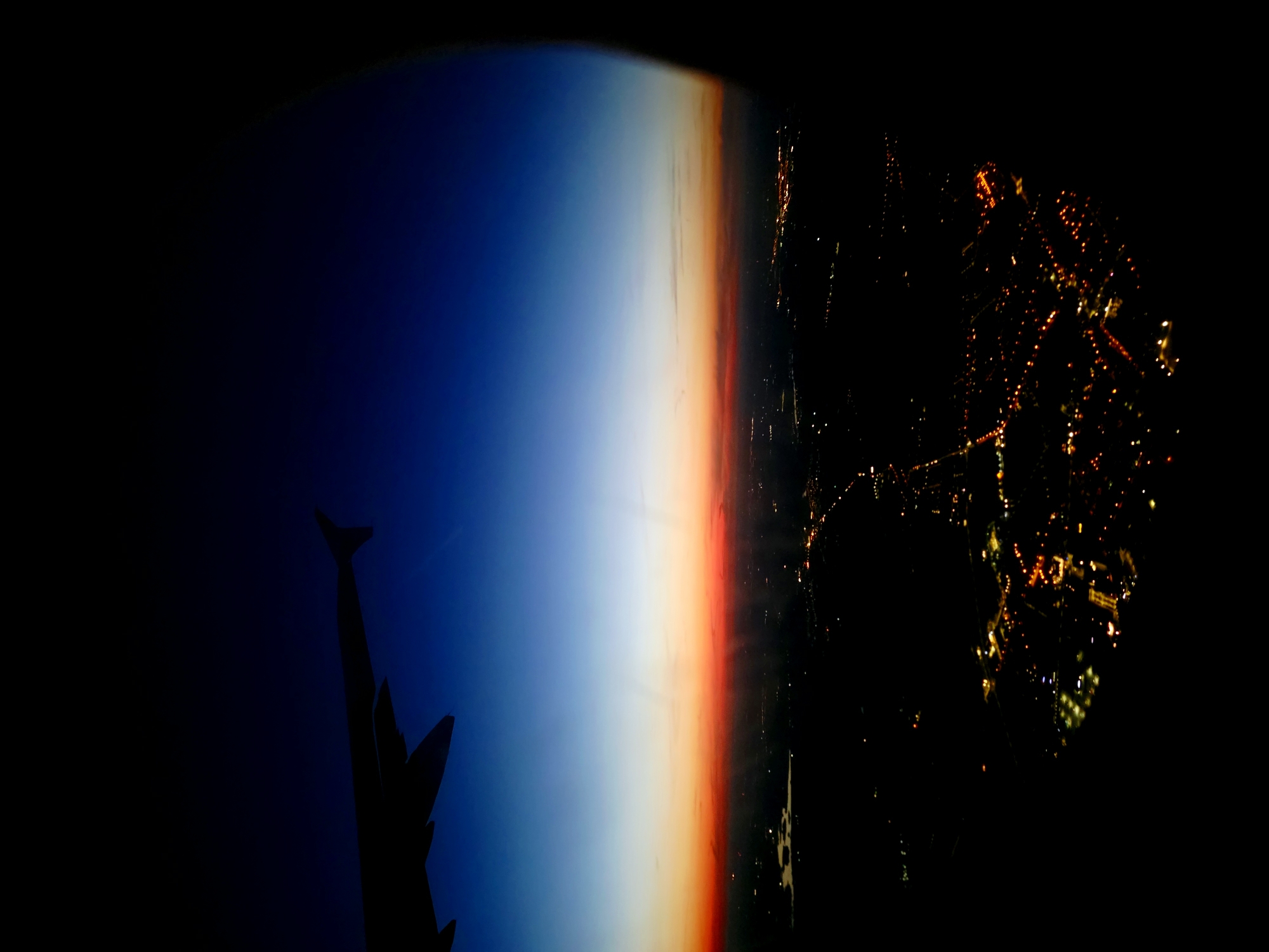 This is a vertical, aerial shot taken from an airplane window at dusk or dawn. The upper portion of the image features a gradient of deep blue sky transitioning to a vibrant band of orange and red at the horizon. The silhouette of an airplane wing is visible in the upper left corner. Below the horizon, the lights of a city are spread out across the dark landscape. The city is identified as Lusówko, Poland. The lights appear as small, scattered points of yellow and white, indicating urban development. Some areas of darker patches suggest water bodies or unlit terrain. The overall atmosphere is serene and captures the transition from day to night, or vice versa, from a high vantage point.