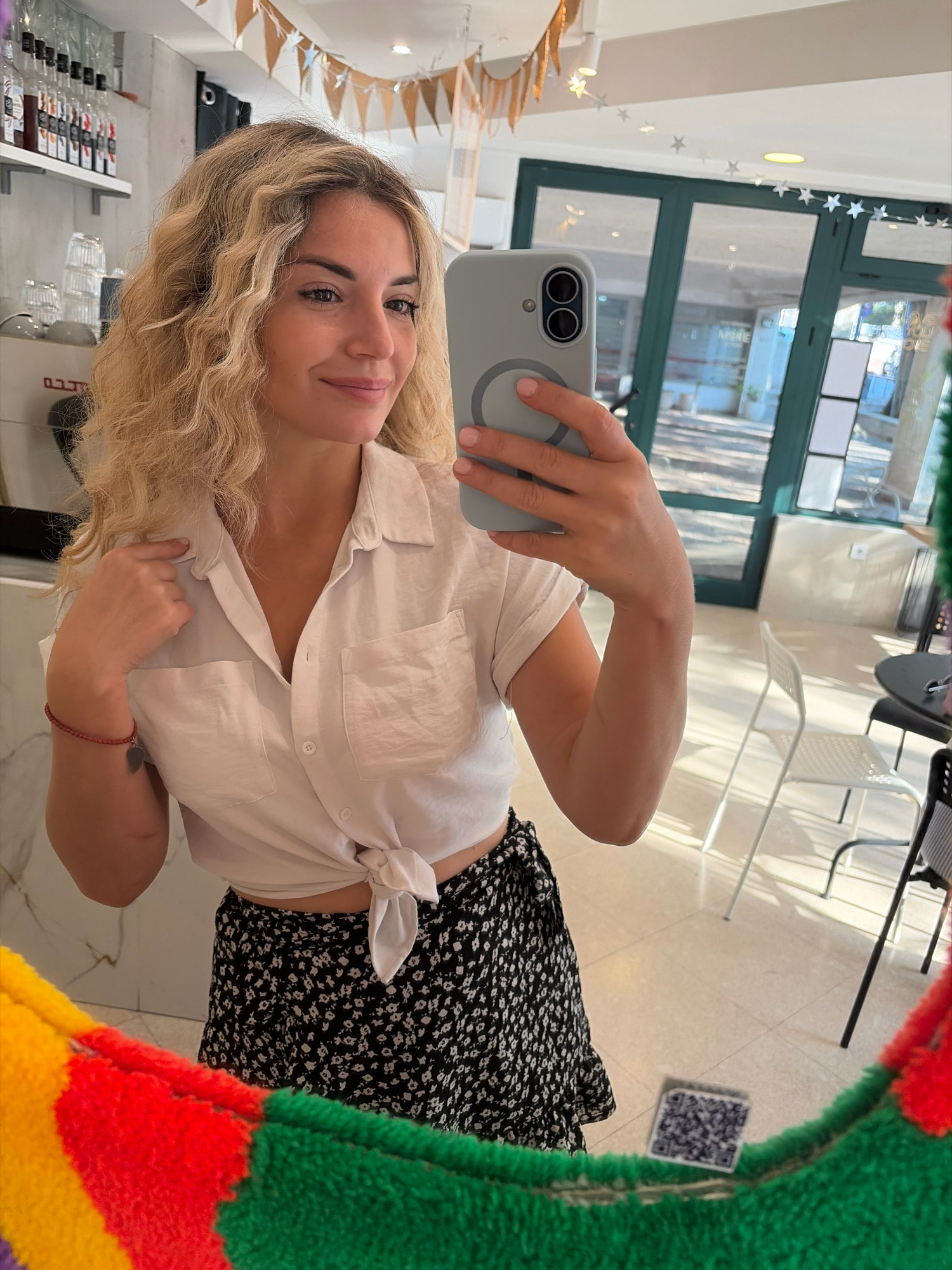 A young woman with blonde, curly hair takes a selfie in a brightly lit indoor setting. She is wearing a white, short-sleeved button-down shirt, tied at the waist, and a black and white floral print skirt. She is holding a light grey phone in her right hand, using it to take the picture in a mirror. The mirror's frame is colorful, appearing to be made of fuzzy, multi-colored fabric bands in red, yellow, green, and purple.
In the background, reflected in the mirror, is a cafe or shop interior. Shelves with various bottles, likely syrups or liquors, are visible on the left. A coffee machine is also present. The environment appears modern and clean, with white walls and light-colored flooring. Large glass doors with dark frames are visible in the background, allowing natural light to enter. Several white chairs and a dark round table are also reflected in the mirror.
The overall impression is that of a casual, possibly vacation, snapshot taken in a trendy establishment in Budva, Montenegro, given the context of the provided image source.