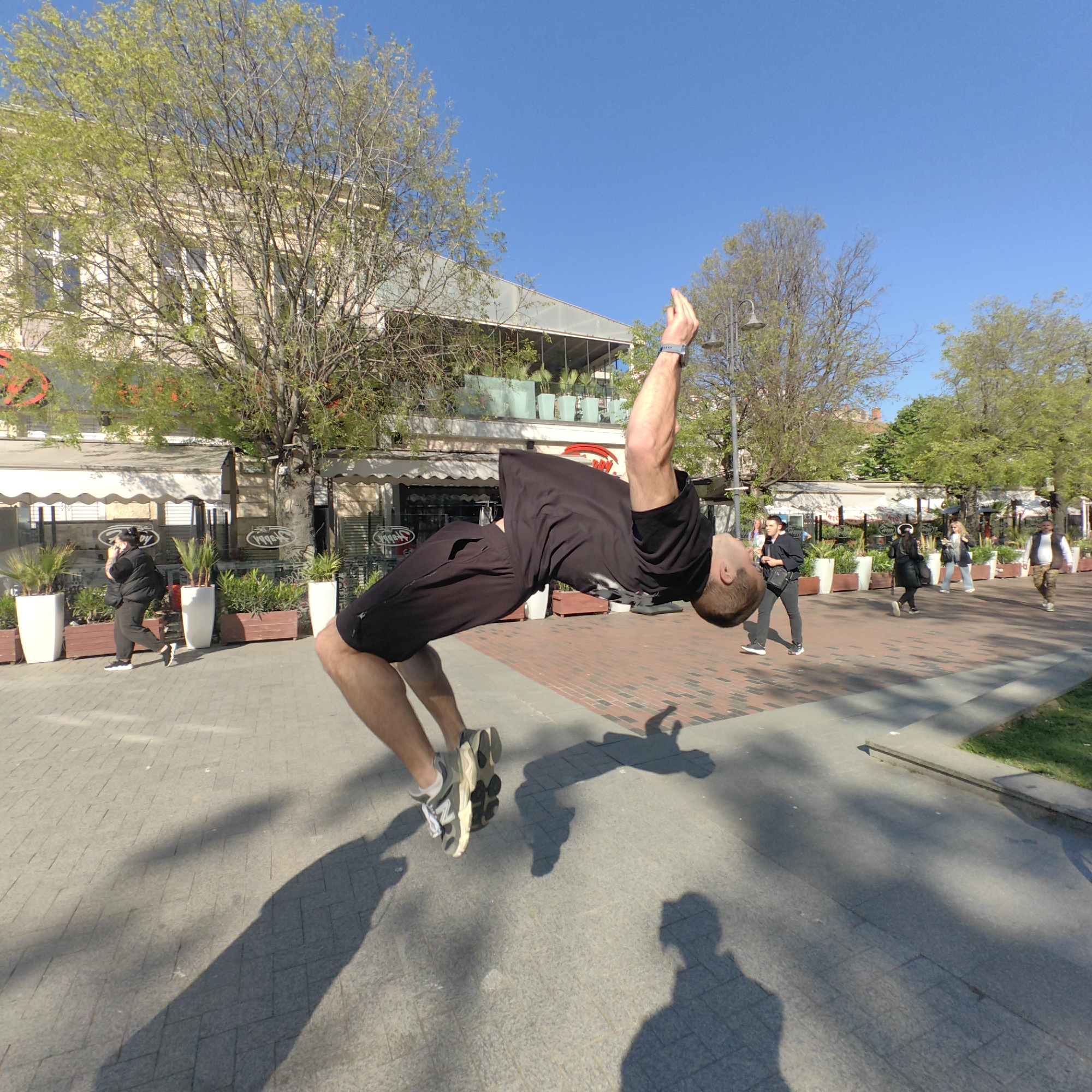 A man is performing a backflip in a public space. He is wearing a black t-shirt, black shorts, and athletic shoes. His arms are raised, and his body is arched backward. He is casting a distinct shadow on the ground.
In the background, there's a building with shops and restaurants along the street, marked by awnings, planters, and outdoor seating. People are walking on the brick-paved street, and the scene is set during the day under a clear blue sky. Trees line the street, adding greenery to the urban environment.
Based on the architectural style and public space characteristics, the location is identifiable as central Varna, Bulgaria, specifically near the pedestrian zone adjacent to the Sea Garden.