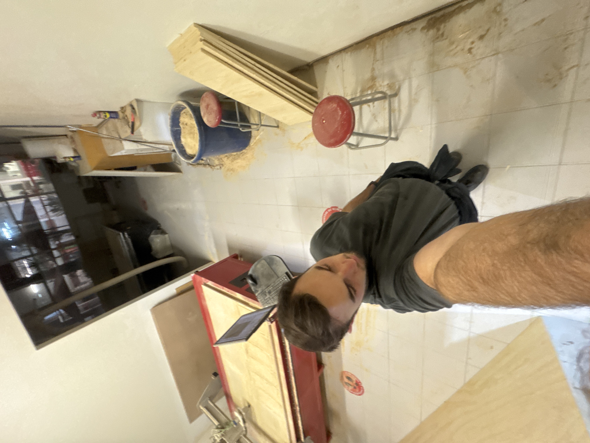 The image is an indoor shot, possibly taken in a workshop or home renovation space. A man is in the foreground, appearing to be taking a selfie. He is wearing a grey t-shirt, black shorts, and black shoes.
Behind the man, a red and wood workbench is visible, with a tablet placed on it. A grey baseball cap also sits on the workbench. To the left, there is a TV. In the background, a stack of plywood and a blue bucket filled with sawdust are apparent, along with red stools and an area with stains or dirt on the tiled floor. The location within San Bernardino Tlaxcalancingo, Mexico, is not discernible from the visual information provided.