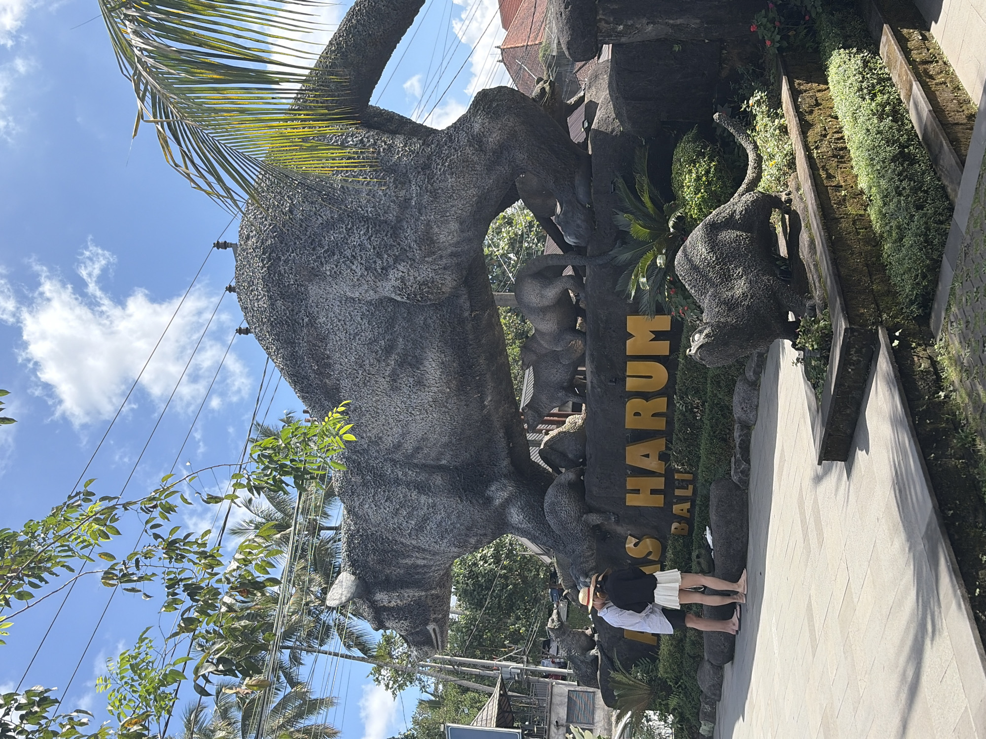 The image shows a large stone sculpture of a creature resembling a bear or a large cat, with smaller sculptures of similar creatures around its base. The sculpture is located in a park-like setting with lush greenery and palm trees. In the foreground, two people, a male and a female, are standing and looking at the sculpture. The male is wearing a white shirt and shorts, and the female is wearing a black top and a white skirt. They are both wearing hats. In front of the sculpture, there is a sign with yellow lettering that appears to read "DAS HARUM BALI". The sky is blue with some white clouds. The overall impression is that of an outdoor tourist attraction. This is the Bali Bird Park in Tegallalang, Indonesia.