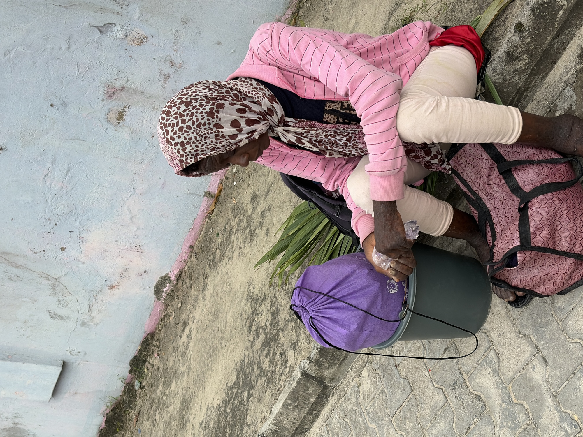 A woman sits on a pavement in Igboefon, Nigeria. She is wearing a pink striped jacket, a dark shirt, light-colored pants, and a headscarf with a brown and white spotted pattern. A purple drawstring bag sits on top of a dark grey bucket next to her. A pink and black duffel bag is on the ground in front of her. There are palm fronds to her left. The pavement is made of interlocking grey stones. A light blue wall with pink trim and visible cracks runs along the back of the scene. The woman is holding a clear plastic bag in her right hand.