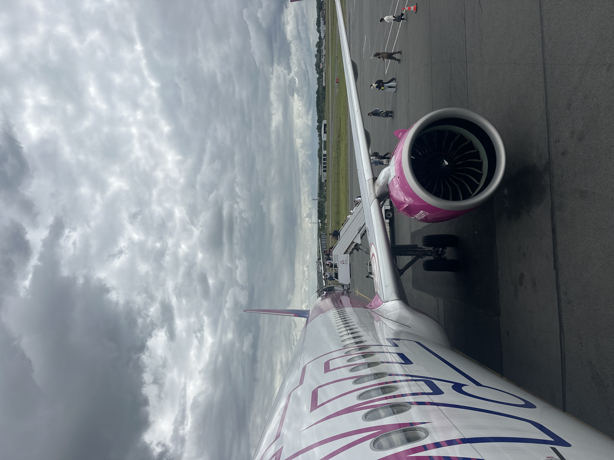 The image, viewed upright, depicts an airport scene from an airplane window, focusing on the aircraft's wing and engine against an overcast sky. The main subject is a Wizz Air airplane, easily identifiable by its distinct pink and purple livery. The word "WIZZ" is prominently displayed in large purple letters along the white fuselage, and "air.com" is visible on the pink engine cowling, which also features the Wizz Air logo.
The sky is dominated by thick, grey clouds, indicating cloudy weather. On the tarmac below, several people are seen walking, suggesting passengers are either boarding or disembarking the aircraft. A set of mobile stairs is connected to the airplane's door, facilitating access. An orange cone is also visible on the tarmac near the wing. In the background, beyond the paved ground, there is a stretch of green land and some distant structures, characteristic of an airport environment. The scene captures a moment of activity on the ground at an airport, likely Beauvais–Tillé Airport in Tillé, France, during daytime.