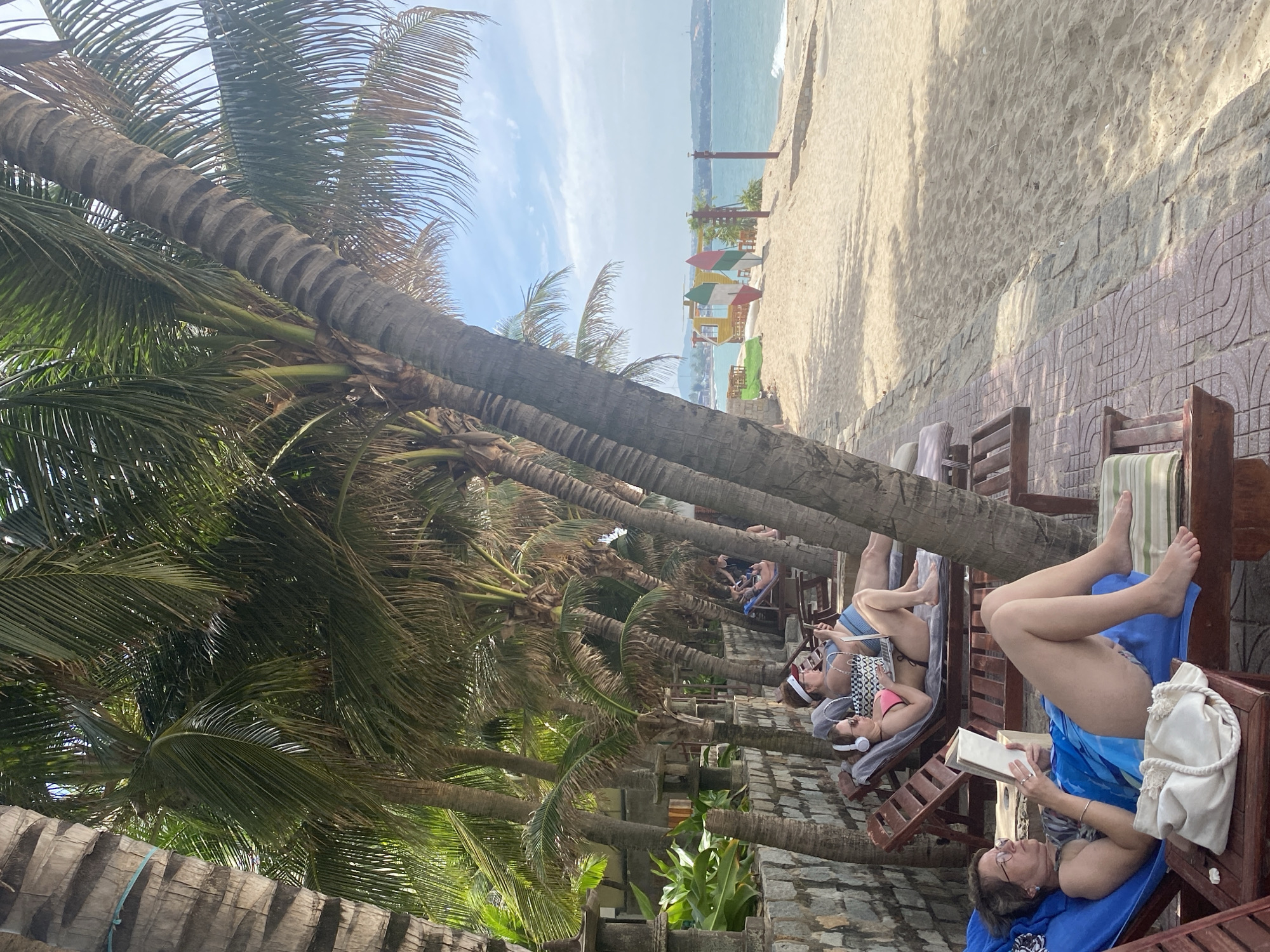 A beach scene in Phu Thuy Ward, Vietnam, shows several people relaxing on lounge chairs under palm trees. In the foreground, a woman with glasses wearing a blue sarong and a dark top is reading a book. To her right, another woman in a pink bikini top and headphones is looking at a phone, with a laptop on the lounge chair beside her. Further back, other individuals are visible on lounge chairs. The sandy beach extends to the ocean, where gentle waves are visible. In the background, a yellow structure with colorful surfboards leans against it stands on the sand. The scene is characterized by bright daylight and a clear sky with some clouds.