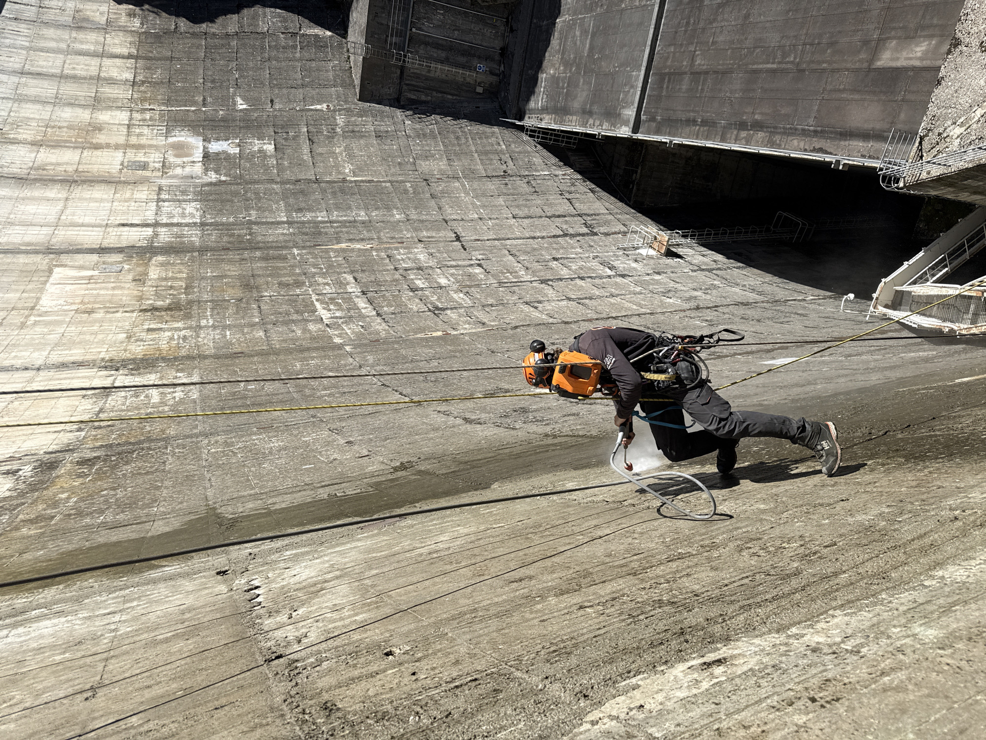 A person in work attire, including a safety harness and hard hat, is rappelling down the face of a large concrete dam. The person appears to be operating a pressure washer, cleaning the dam's surface. The location is likely the Castillon Dam, near the city of Castellane, France, based on the architecture and environment depicted. The dam's concrete surface shows varying degrees of staining and discoloration. Ladders and scaffolding are visible near the top of the dam. A yellow rope spans across the surface.
