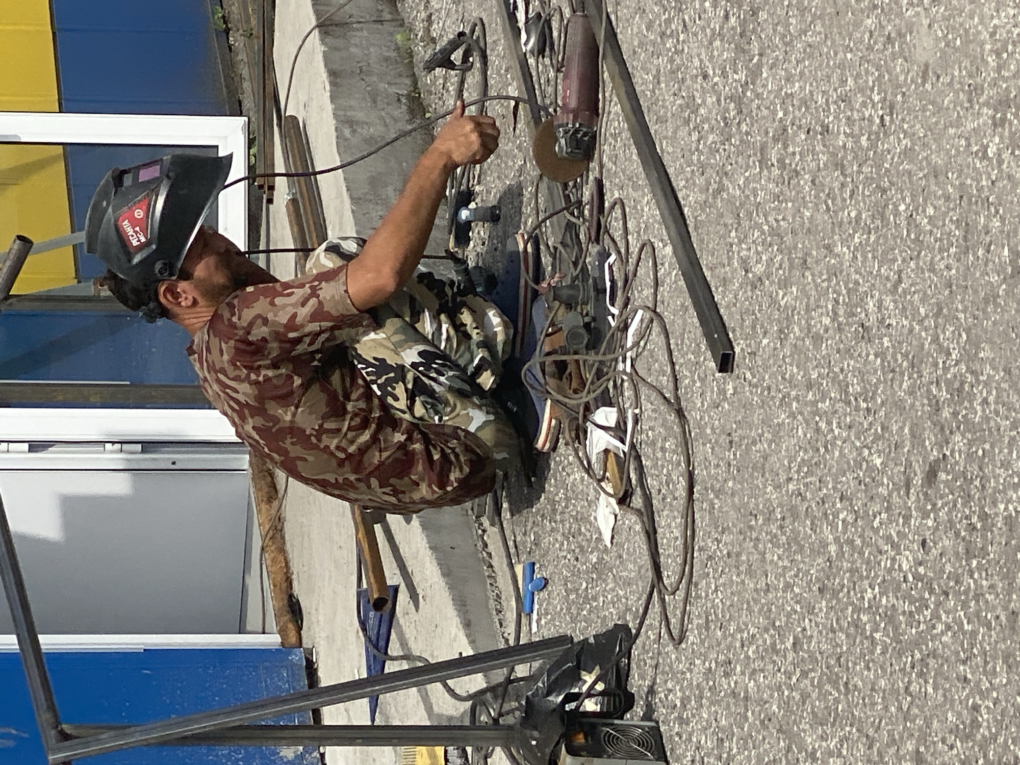 A male welder, wearing a camouflage shirt and pants, and a black welding helmet, is crouched on a concrete surface outdoors. He is holding a welding electrode and appears to be in the process of welding metal. Various tools and equipment are scattered around him on the ground, including an angle grinder, electrical cords, and metal pipes. In the background, there is a building with blue and yellow sections and a white door or window frame. The scene suggests an outdoor construction or repair site. This image was taken in Novokuznetsk, Russia.