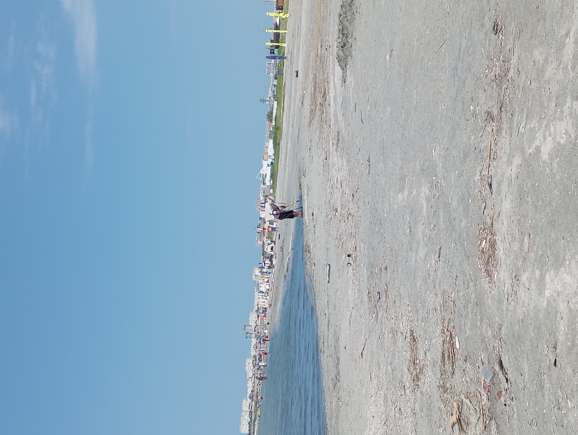 This is a medium-distance, eye-level, wide shot of a beach on a clear, sunny day in Ömirzaq, Kazakhstan. The ocean is on the left, and the beach stretches to the right. In the distance, there is a city skyline with several buildings. On the beach, there are people scattered around, some near the water and others further back. A few tents and tall, brightly colored poles are also visible. In the foreground, a person is raking the sand, and there is some debris on the beach. The sky is a clear blue with a few wispy clouds.
