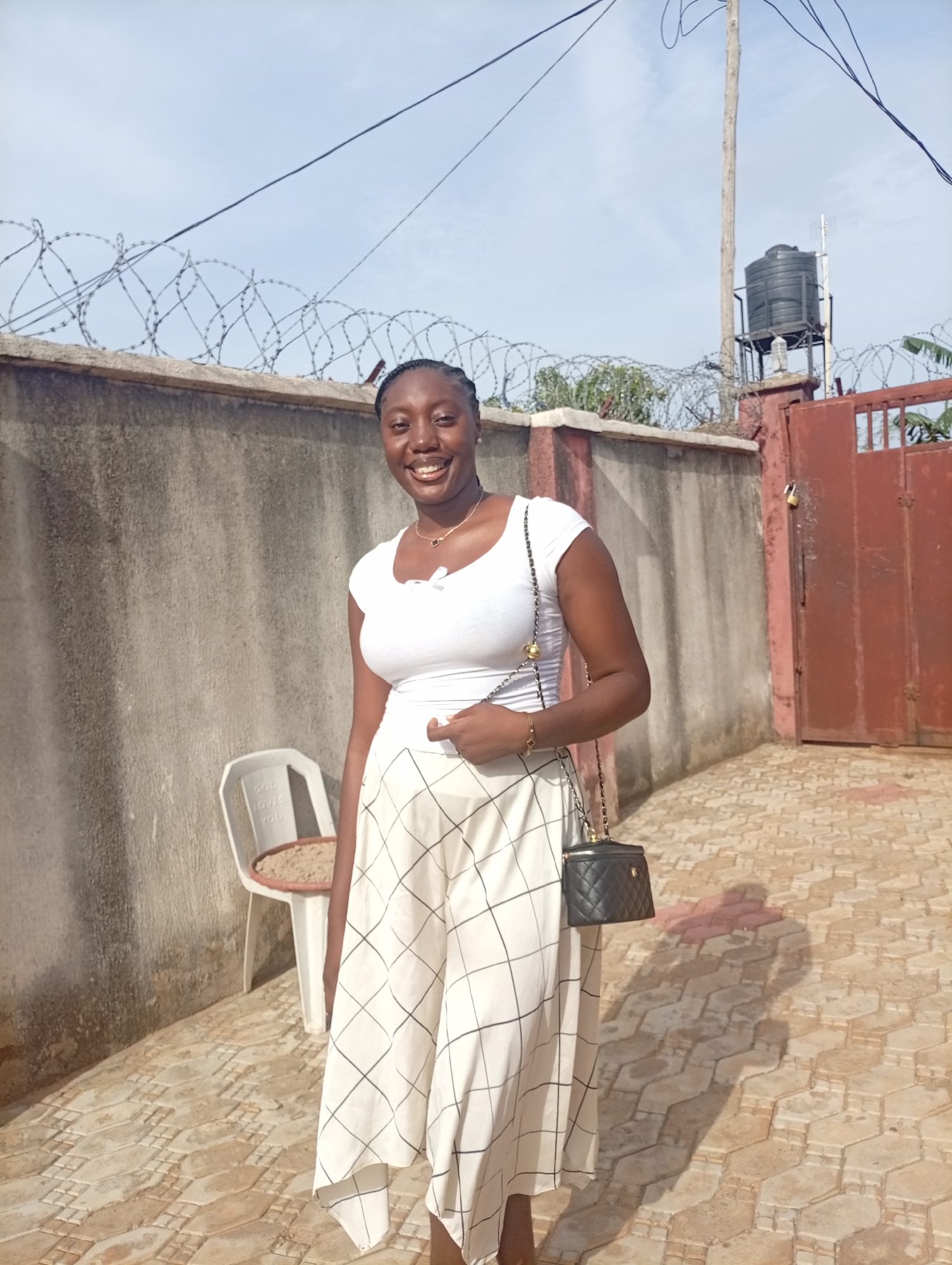 A young woman stands in a courtyard paved with tan hexagonal stones. She wears a white t-shirt, a long white skirt with black gridlines, and a black purse on a chain strap. Behind her, a concrete wall topped with barbed wire runs along the left side of the frame. To the right of the wall, a red metal gate is visible. Beyond the gate, a water tank on a metal stand sits on top of a pole. A white plastic chair with writing on the back is located beside the wall. The location is the city of Unknown, Unknown.