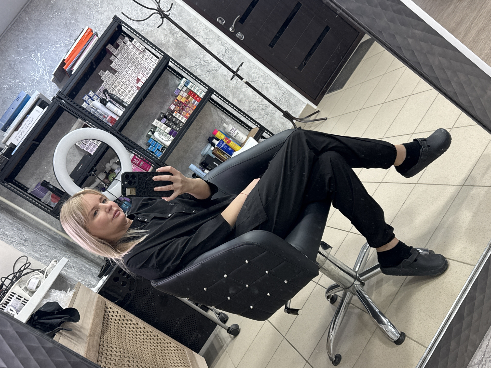Here's a detailed description of the image:
**General Scene:**
The image is a selfie taken in what appears to be a hair salon or stylist's studio. The location is possibly in the city of Chaikovsky, Russia. The shot is angled, showing the environment through a mirror.
**People:**
* A woman is visible, seated in a salon chair. She has blonde hair and is wearing black clothing and black shoes with black socks. She's holding a smartphone, presumably taking the selfie.
**Objects:**
* **Salon Equipment:** A stylist's chair with a studded design, shelves stocked with hair coloring products, a ring light, and what seems to be a hair dryer or steamer on the lower left.
* **Mirror:** The shot is taken in a mirror, the frame of which is visible.
* **Door:** A dark-colored door with silver hardware is present in the background.
* **Phone:** A phone with leopard print is taking the photo.
**Actions and Interactions:**
* The woman is taking a selfie in the mirror.
* She is seated in the stylist's chair.
**Environment:**
* The interior appears to be a salon or studio.
* The walls are a light color with a marbled or textured finish.
* The floor is tiled.
* The area seems organized with shelves holding hair products and equipment.
