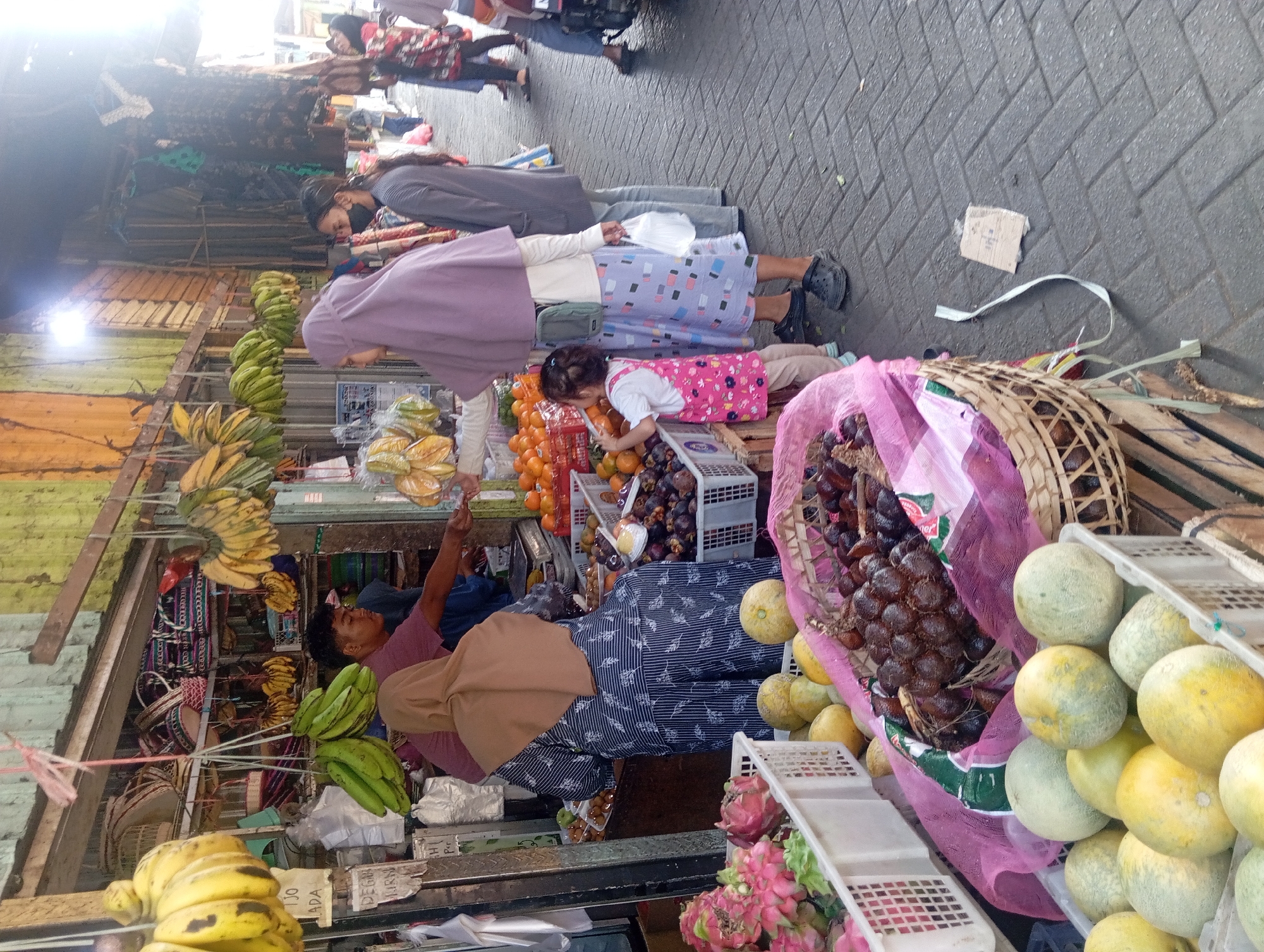 An outdoor market scene in Kepulungan, Indonesia, features a fruit stall with a variety of fresh produce. In the foreground, several baskets are filled with fruits including dragon fruit, melons, and salak (snake fruit). Behind these, a woman in a blue and white striped dress and a light brown hijab is seen from the back, interacting with a male vendor. She is holding a light pink scarf. To her left, a young child in a pink dress is reaching towards a basket of fruit. Further back, other shoppers and vendors are visible amidst the stalls, with a variety of goods displayed, including bananas, oranges, and woven baskets. The ground is paved with brickwork. The market appears to be a busy, open-air environment.