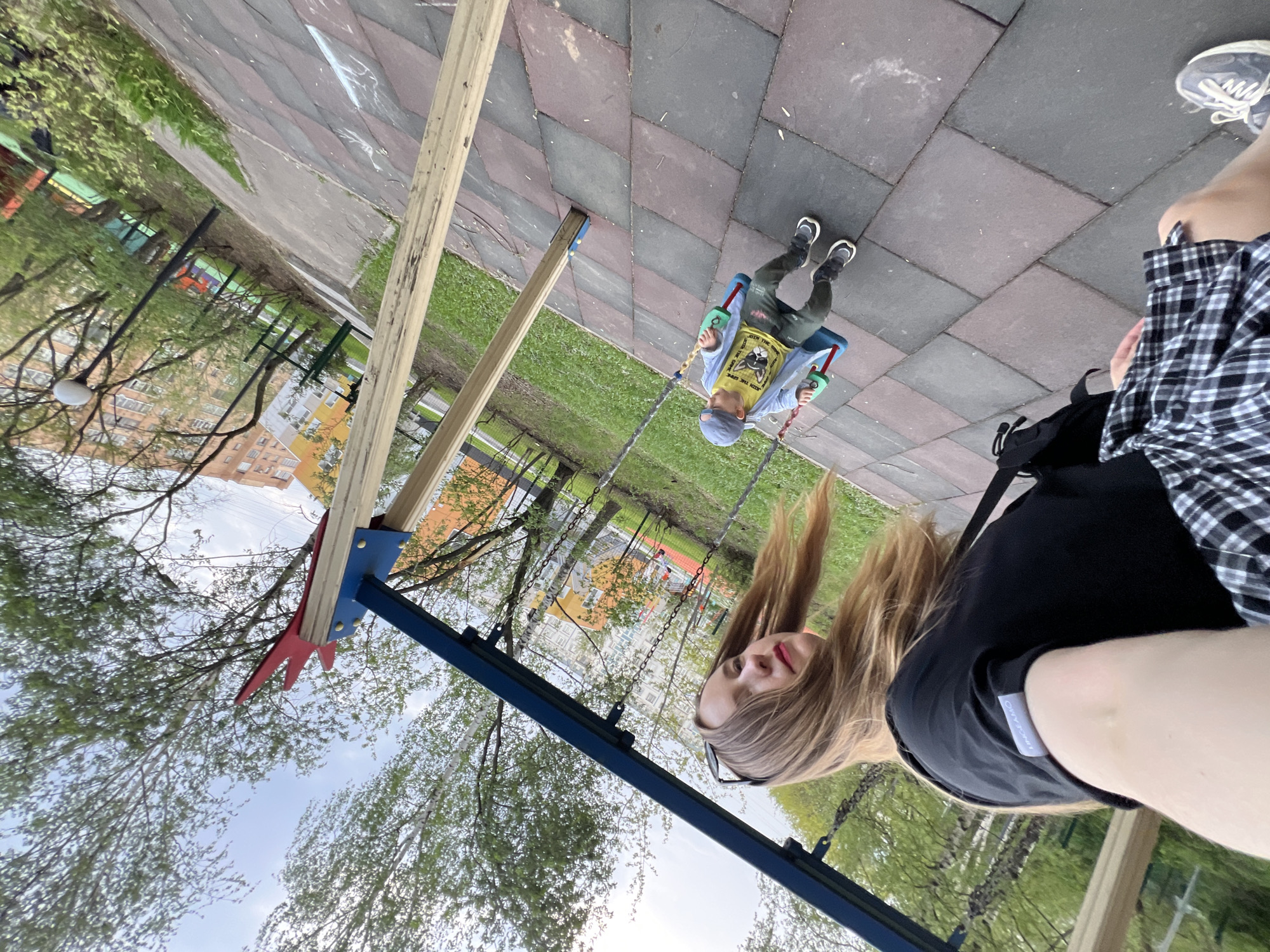 A selfie was taken at a playground in Mytishchi, Russia. A young child is swinging on a swing. The child is wearing a blue hat, blue jacket, and green pants. A woman with long blonde hair is taking the picture. She is wearing a black t-shirt and a checkered shirt. The playground has a rubber surface. Trees and buildings are visible in the background.