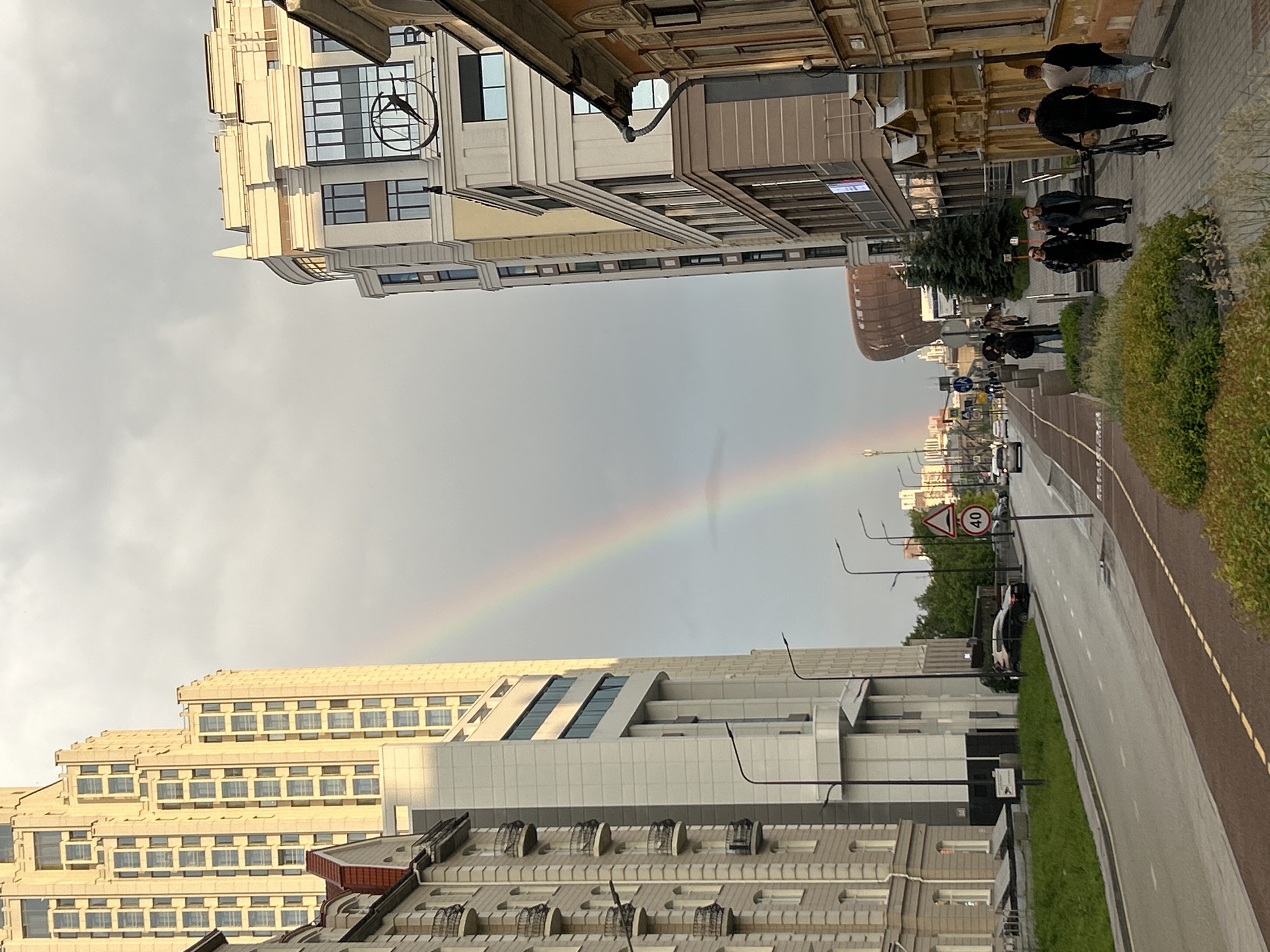 The image depicts an urban street scene in Yekaterinburg, Russia, specifically on Boris Yeltsin Street, with a rainbow visible in the sky. The sky is largely overcast, yet a distinct rainbow arcs from the mid-left towards the mid-right in the upper half of the frame.
On the left side of the street, a tall, multi-story modern skyscraper with a stepped facade and numerous rectangular windows, identified as the Demidov Plaza, dominates the left background. Below and slightly in front of it, a lighter-toned building with multiple balconies and sections of a reddish-brown tiled roof is visible. Streetlights with multiple arms are positioned along the street. The road features two lanes, a green grassy median strip, and a reddish-brown dedicated bicycle lane on the left side, which runs parallel to a pedestrian sidewalk. A black sedan is parked on the street to the left. A triangular warning sign and a circular speed limit sign indicating "40" km/h are visible on a pole near the street.
On the right side of the street, another multi-story modern building is present, featuring a large, stylized clock face with an abstract bird-like hand design near its upper portion. This building is part of the Yeltsin Center complex. Adjacent to it on the right, older, ornate buildings with yellowish-orange facades and decorative architectural elements line the street. Several pedestrians are walking on the paved sidewalk on the right. A group of four individuals is walking towards the viewer in the mid-ground on the right sidewalk; one person is carrying a bicycle. Further down the street, a distinctive large, rounded building with a copper-colored facade, identified as the main structure of the Boris Yeltsin Presidential Center, is visible in the background. Scattered greenery and bushes line the sidewalks and medians.