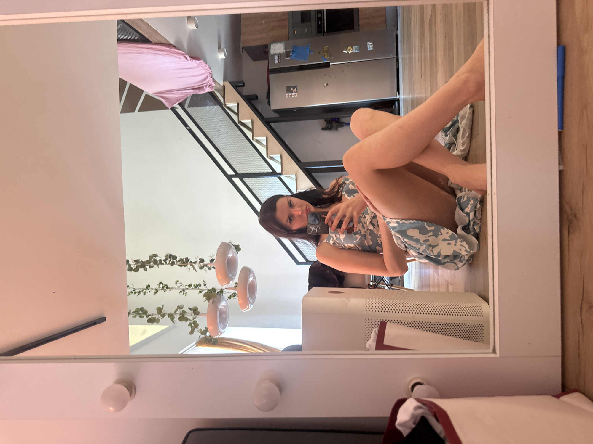 A woman is taking a mirror selfie while sitting on the floor in front of a full-length mirror. She is wearing a light-colored, patterned dress and has her legs crossed. She is holding a smartphone in her right hand, and her left hand is resting on her left knee. The mirror reflects a room with a staircase, a refrigerator, and a kitchen area. There is a white air purifier to her left, and a hamper with clothes to her far left. The floor is wooden, and the walls are painted a light color. The lighting in the room is soft, with some pinkish tones.