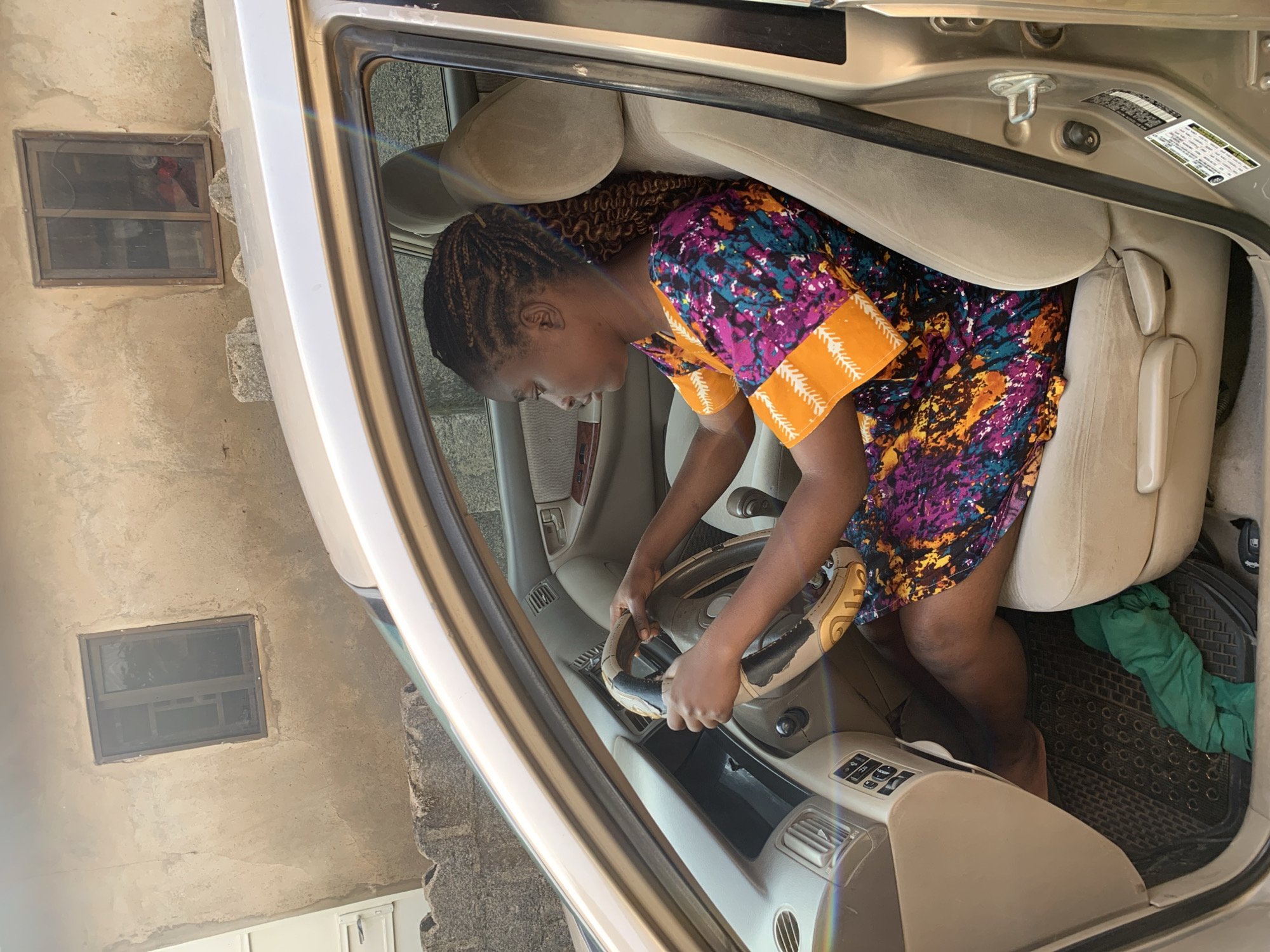 A woman is shown sitting in the driver's seat of a car, with her hands on the steering wheel. She is wearing a colorful dress and her hair is braided. The car's interior appears to be light beige, and there is a black floor mat on the driver's side with a green cloth lying on it. The exterior of the car is a light gold color. The background shows a rough, tan-colored wall with two small windows and some rough stone structures. The scene suggests a setting in Sabon Tasha, Nigeria, possibly a residential area given the wall and windows.