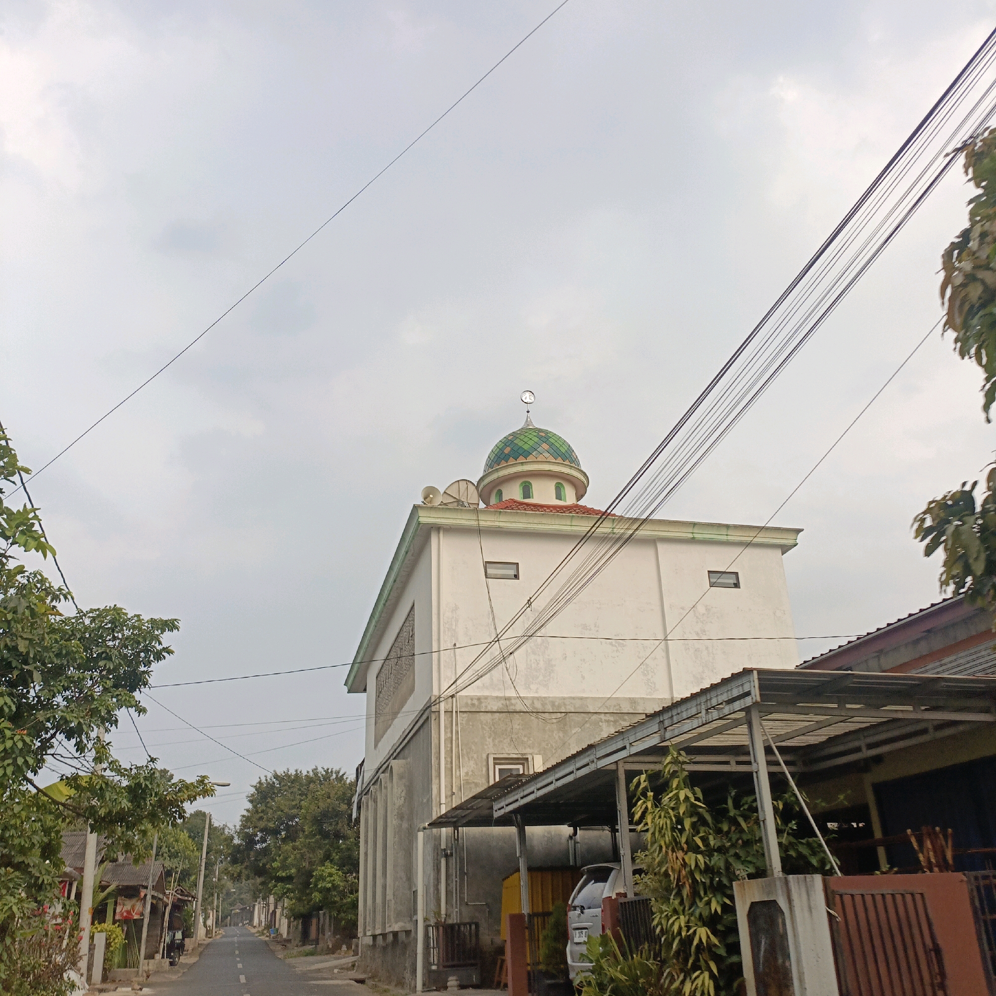 The image depicts a street scene in Salatiga, Indonesia, featuring a white building with a green-domed roof, likely a mosque, as indicated by the crescent moon symbol on top. The building has two visible windows on the upper level, and a smaller rectangular window on the side. In front of the building, there is a covered parking area with a silver vehicle parked underneath, partially obscured by foliage. A brown metal fence is also visible. The street is asphalted and appears to be a quiet residential road, with trees and smaller houses lining either side. Power lines are strung across the sky, adding to the urban overhead infrastructure. The sky is overcast, suggesting a cloudy or hazy day.