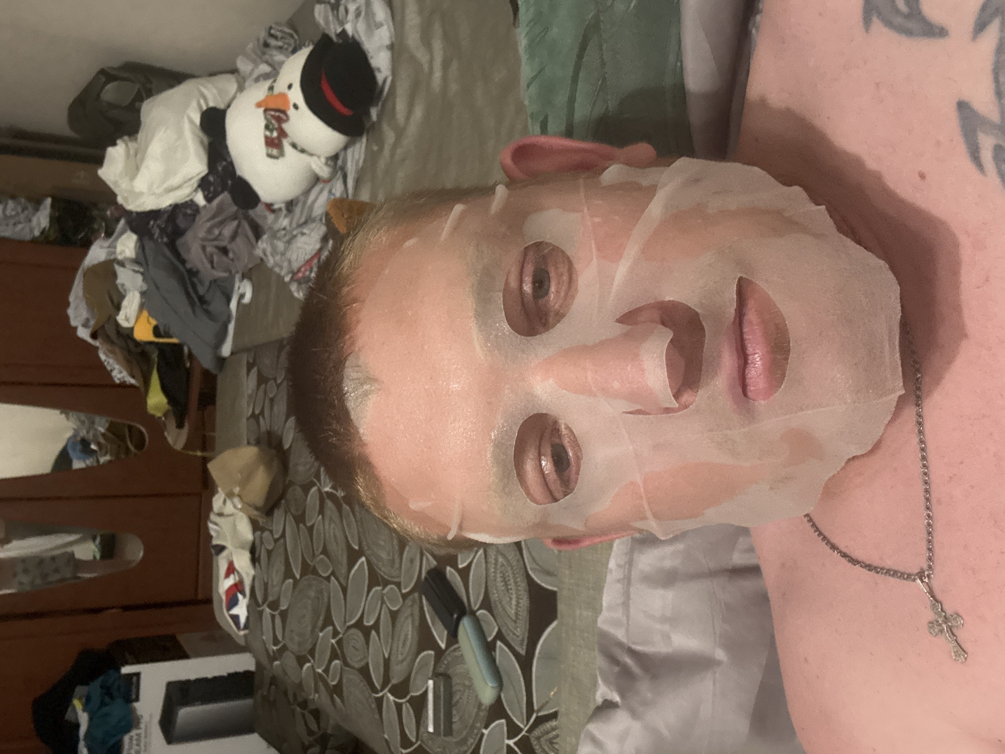 The image shows a close-up, high-angle shot of a man with a sheet mask on his face. He is lying down, with his head tilted slightly. The man has short, light brown hair and is wearing a silver chain with a cross pendant. His skin appears pinkish and slightly sweaty. The background of the image is a messy bed with rumpled sheets and several items of clothing scattered around. There is a stuffed snowman toy lying on the bed to the right. To the left, a portion of a brown wooden wardrobe with mirrors is visible, along with a cardboard box. The lighting is dim and appears to be artificial. The setting is an indoor environment, likely a bedroom.