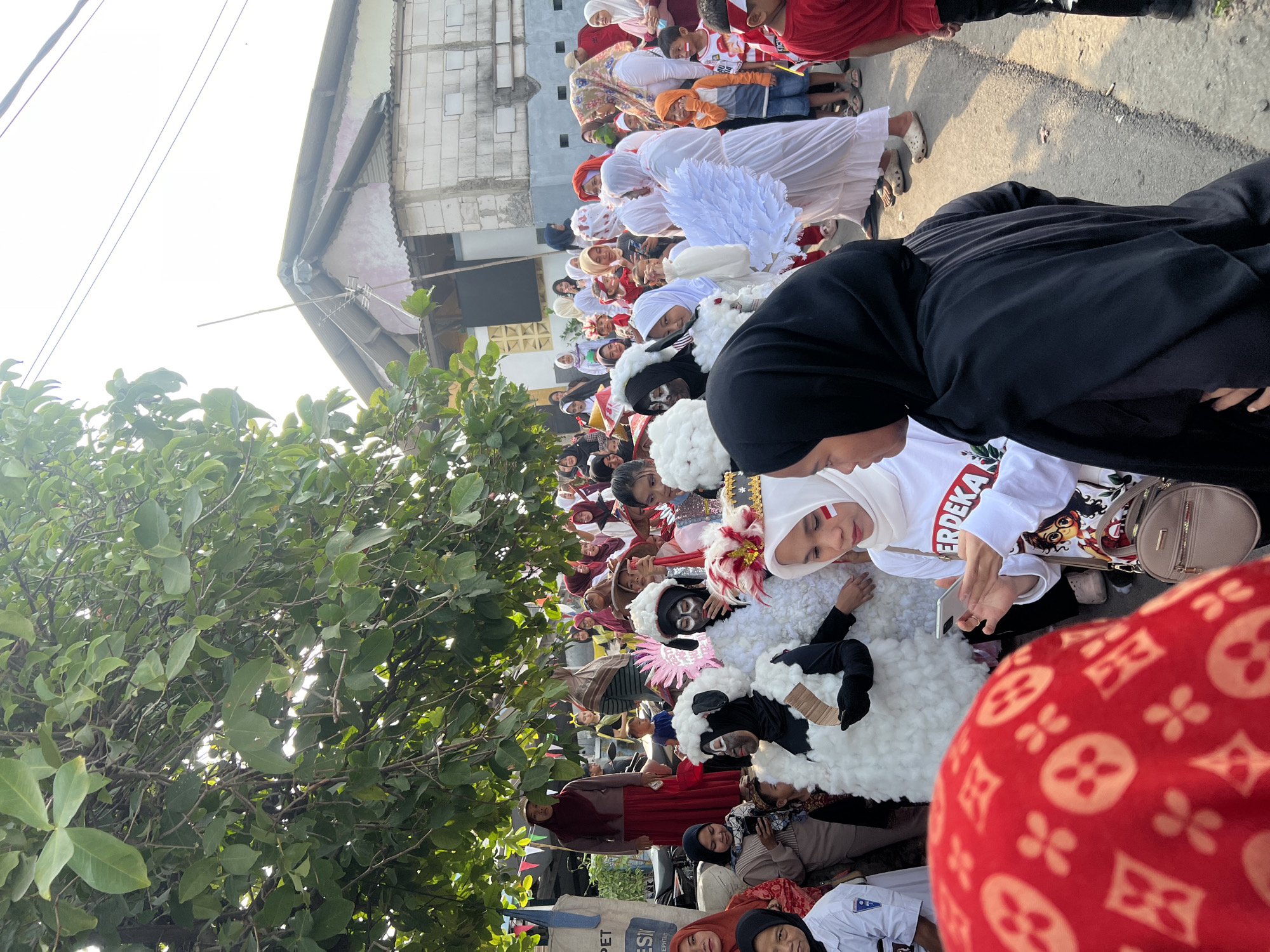 This is a medium shot of a street parade in Klapanunggal, Indonesia, likely celebrating a national holiday given the red and white motifs and the word "MERDEKA" (meaning independence) visible on clothing. A group of people, mostly women and children, are gathered on a paved street. In the foreground, a person wearing a red headscarf with a repeating circular pattern is partially visible. Next to them, a woman in a white headscarf with a red and white stripe on her cheek is looking at her phone. She is wearing a white t-shirt with "MERDEKA" written on it and an illustration of a cartoon character.
Behind these figures, several people are dressed in costumes. Two individuals are adorned in white, fluffy costumes resembling sheep, with black faces and ears. Another person is wearing a white costume with large white wings, possibly representing an angel. Other participants are dressed in a variety of clothing, including traditional headscarves and casual wear. Some children are also present, some holding small Indonesian flags.
The environment suggests a residential area, with buildings visible in the background. A large tree with green leaves overhangs the scene from the left, providing shade. Colorful triangular flags are strung across the street. The lighting indicates it is daytime, possibly late afternoon, with sunlight casting shadows. The overall atmosphere appears festive and communal.