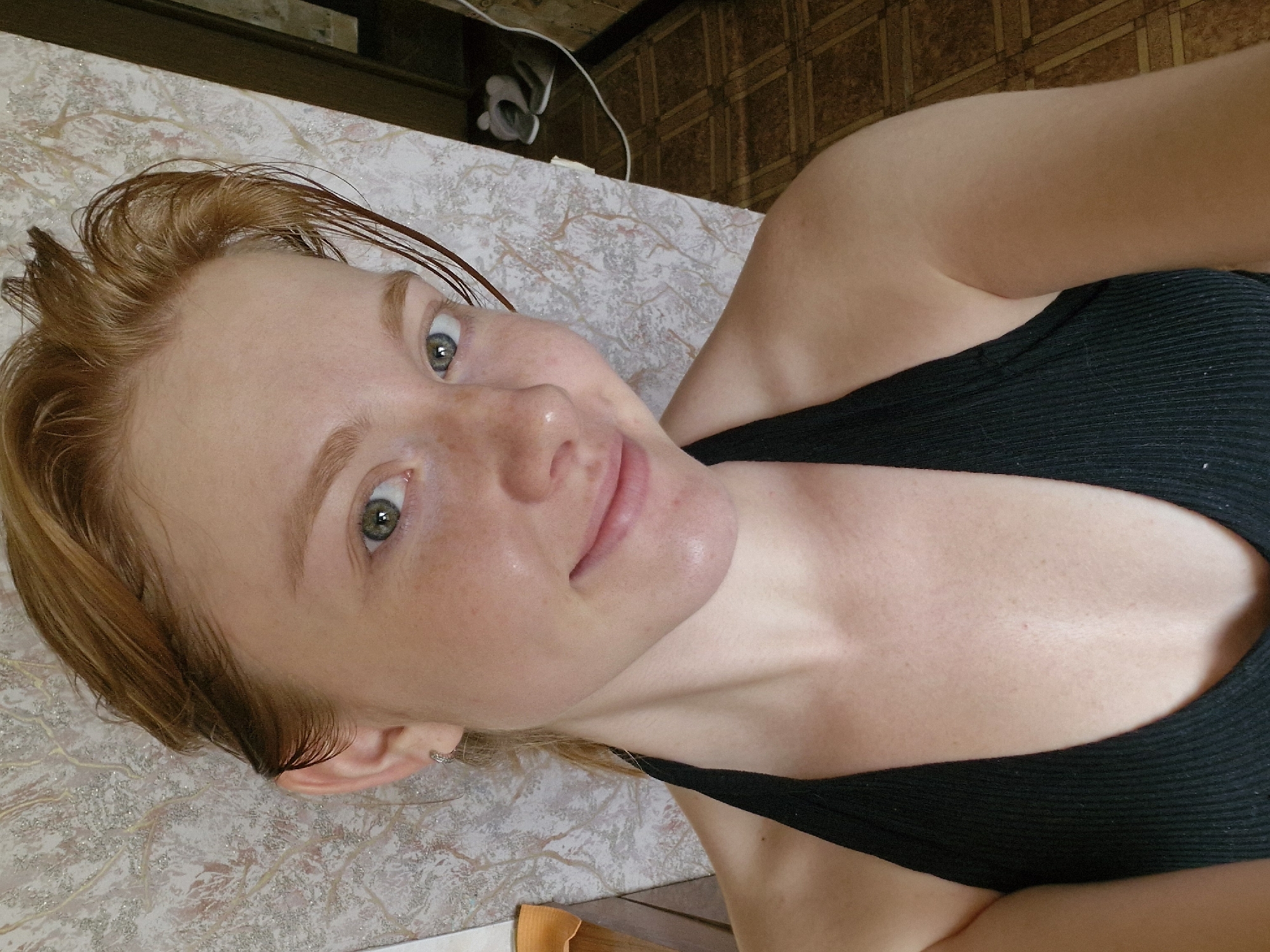 The media file is a close-up, upward-angle shot of a young woman with light skin and freckles, and blonde hair. She is lying down on a surface with a textured, light-colored covering. The woman is wearing a black tank top. Her eyes are open and looking towards the camera. Her hair is slightly damp and styled forward. A small hoop earring is visible in her left ear.
In the background, on a dark surface, there are some objects, including a white cable and what appears to be a shoe or footwear. The flooring in the background has a tiled pattern.
The lighting suggests it might be daytime, possibly indoors, given the presence of indoor flooring and objects. There are no specific cues about the weather. The woman's expression is neutral, and the overall emotion conveyed is casual and relaxed. No text is visible in the media file. The location context is Yekaterinburg, Russia.