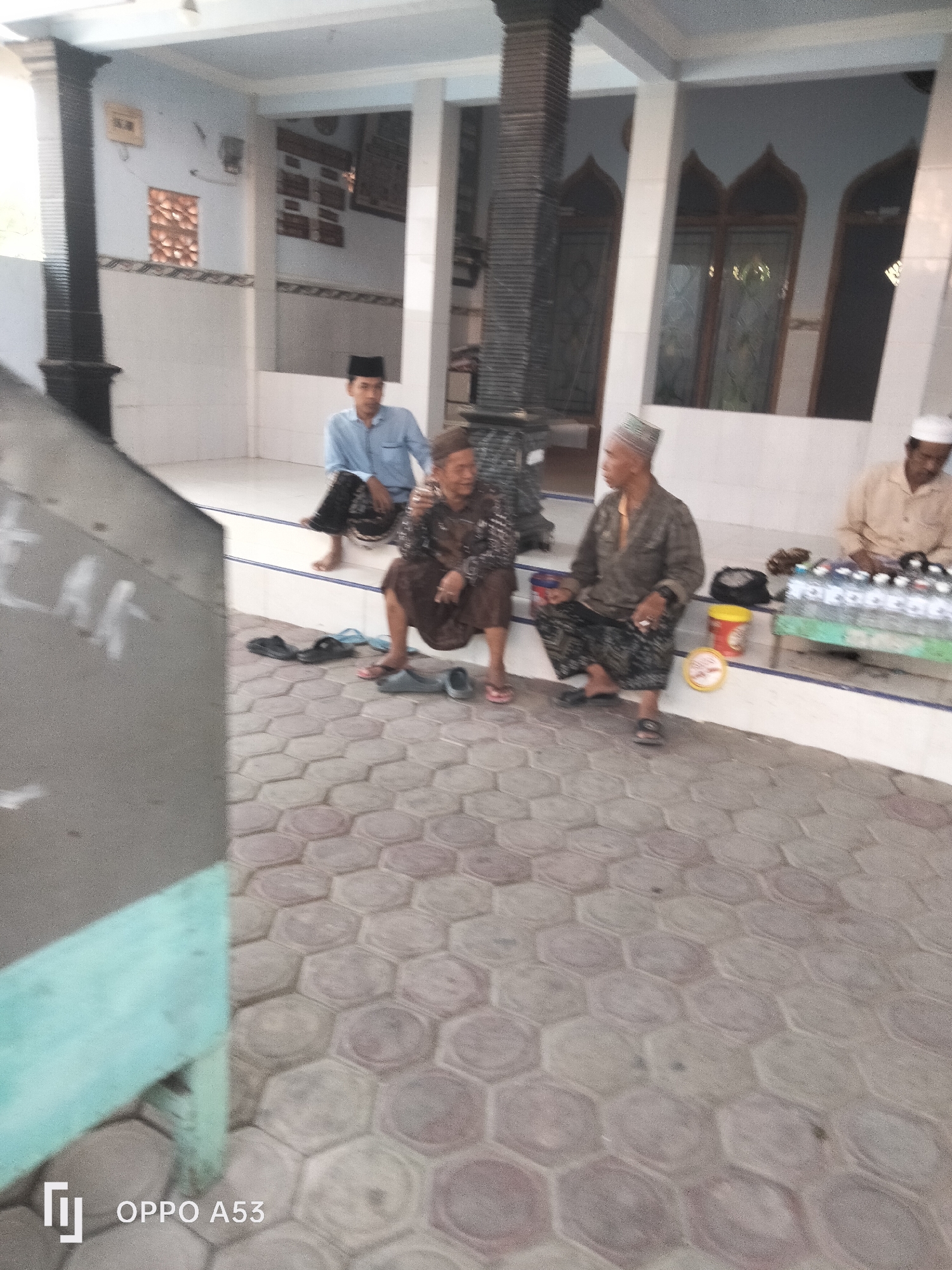 In Badung, Indonesia, four individuals are seated on steps in front of a building with a tiled floor. The building features architectural elements such as columns and arched windows.
On the left, a young man in a light blue collared shirt and dark pants sits with his legs crossed.
To his right, an elderly woman in a dark patterned garment sits with her legs extended. A pair of grey flip-flops is near her feet.
Next to the woman, an older man in a patterned cap and a dark, long-sleeved shirt is seated, holding a cigarette. He is wearing patterned pants and dark sandals. To his right are a red bucket and a yellow circular object.
Further to the right, another man wearing a white cap and a light-colored long-sleeved shirt is seated. In front of him are several bottles of water arranged on a green surface.
The ground in the foreground is paved with hexagonal tiles in varying shades of grey and pink. The environment appears to be an outdoor public space, possibly a religious or community gathering area.
