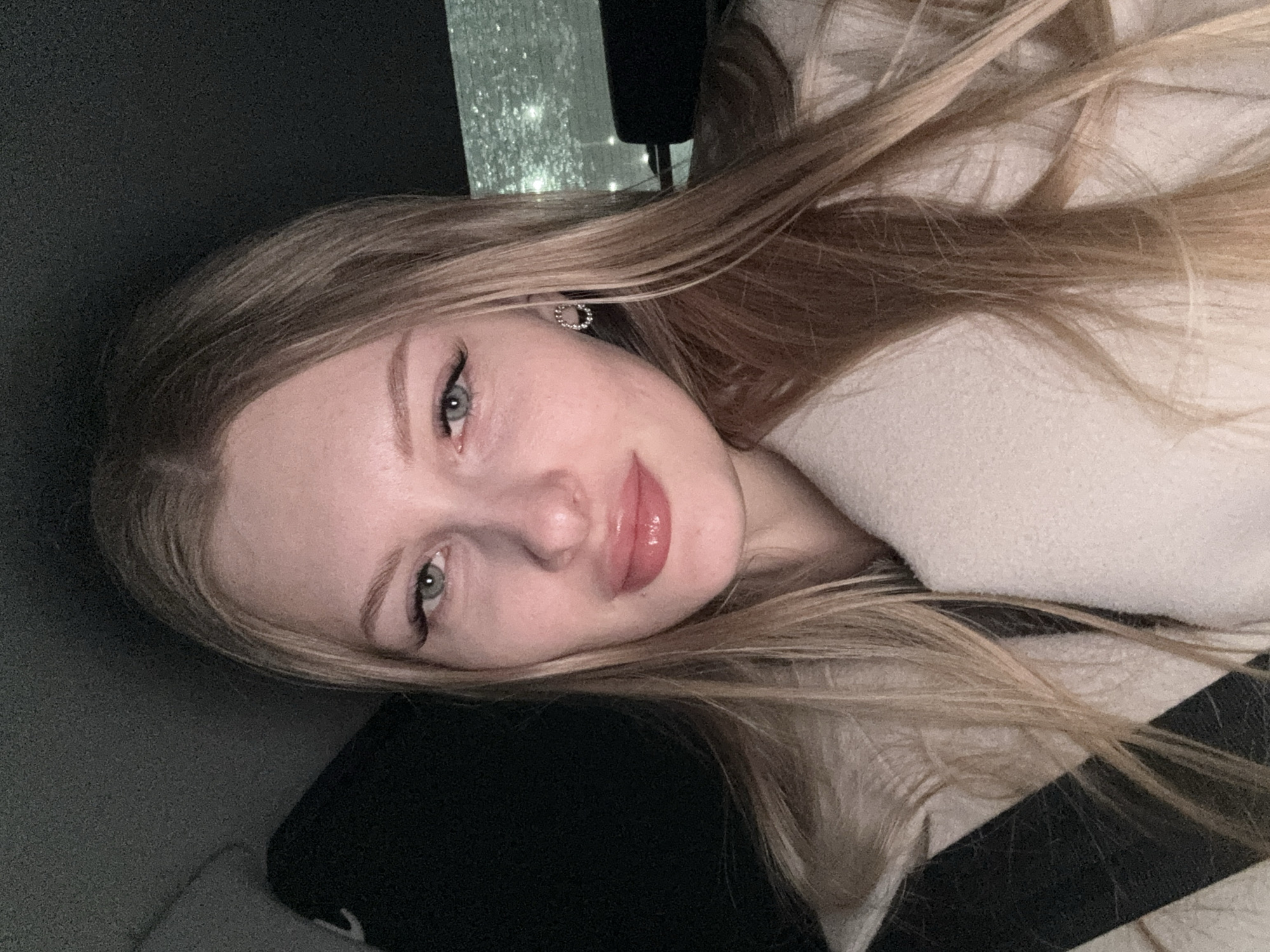 A young adult female is depicted in a close-up, chest-up shot, seated within the passenger compartment of a vehicle in the city of Кондратово, Russia. She possesses long, straight, blonde hair parted to her left, extending over her shoulders. Her face, with a pale complexion, features light-colored eyes (appearing greenish-blue), defined eyebrows, prominent eyeliner, and artificial eyelashes on the upper lids. Her lips are full and appear to be enhanced with a pink, glossy substance. She wears a light-colored, possibly off-white, textured coat or jacket, beneath which a dark garment is visible. A black seatbelt strap crosses her left shoulder and chest. A small, metallic, circular earring, possibly adorned with stones, is present on her right earlobe. The background is dimly lit, revealing dark vehicle interior elements and a rear or side window behind her right shoulder, displaying water droplets or streaks on the glass surface and blurred external lights. The lighting on her face suggests a direct flash or internal light source.