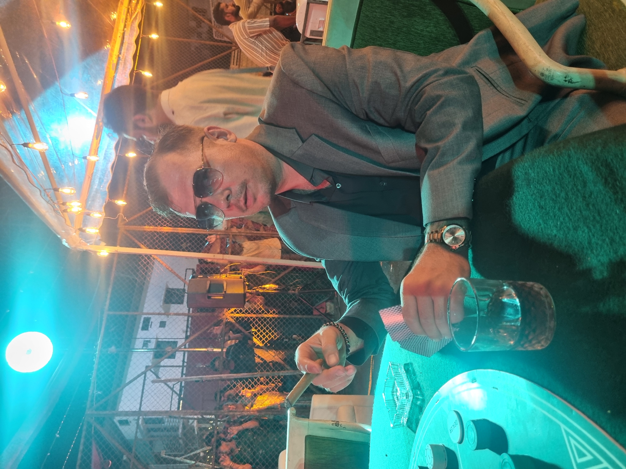 The image displays an evening or nighttime scene, likely outdoors or in a semi-enclosed venue, characterized by artificial lighting. A man in the foreground is the primary subject, captured from the chest up and oriented diagonally due to the original image's rotation. He is wearing a gray suit jacket over a black collared shirt and dark sunglasses. On his left wrist, he has a metallic watch, and on his right, a beaded bracelet. He holds a lit cigar in his right hand and a f fan of playing cards in his left. A clear glass containing a beverage rests on the green felt table before him. The table is equipped with white poker chips or game pieces, and a prominent circular marking on its surface clearly reads "DEALER".
In the background, several other blurred individuals are visible, some standing and others seated, suggesting a social gathering. One person in the mid-ground wears a striped shirt. The setting features metal fencing or scaffolding, contributing to an informal atmosphere. String lights hang overhead, emitting a warm glow, while a large, intense blue light source illuminates the left side of the scene. A light-colored wooden object, possibly a decorative horn or stick, lies on a green surface in the upper right. The overall impression is that of a lively event, possibly a card game like poker, taking place under artificial lights. The man's expression appears neutral and focused, consistent with concentration on a game.
