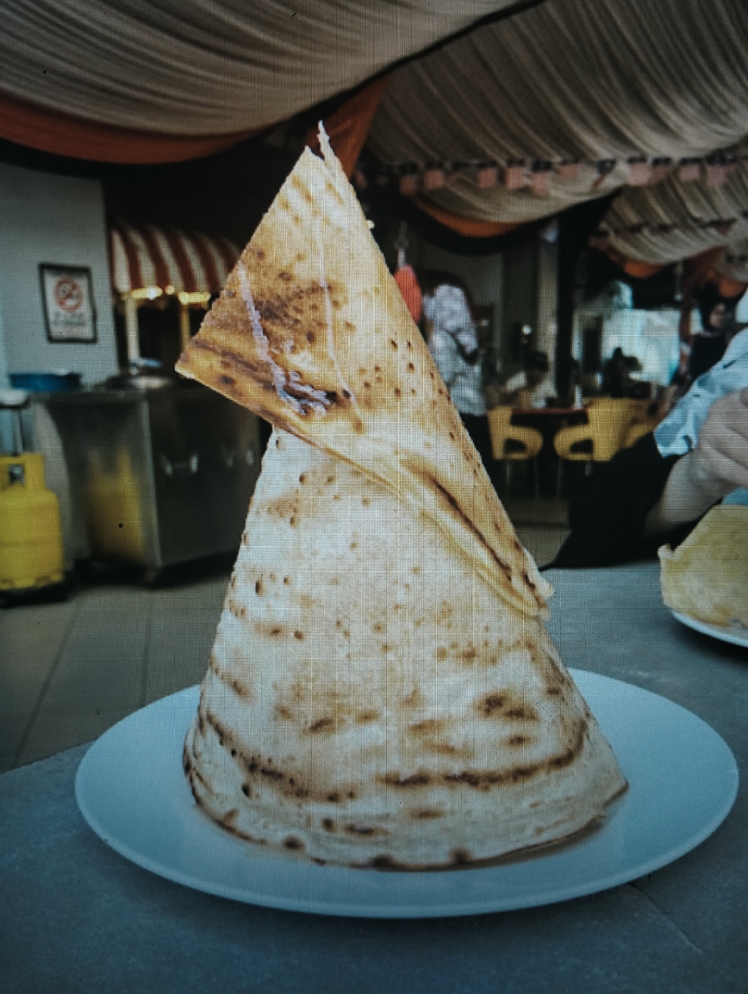 A close-up, low-angle shot depicts a cone-shaped stack of Roti Canai on a white plate, set on a gray table. The Roti Canai is golden brown with darker, burnt-like spots and appears to be made of thin, layered dough. In the background, there is an indoor setting with draped fabric overhead, a yellow propane tank to the left, and blurred figures of people seated at tables with yellow chairs. The overall impression is that of a casual dining establishment, possibly a hawker stall or restaurant, within the city of Medan, Indonesia, known for its street food culture. The lighting is dim, with some areas illuminated by a bright overhead light.