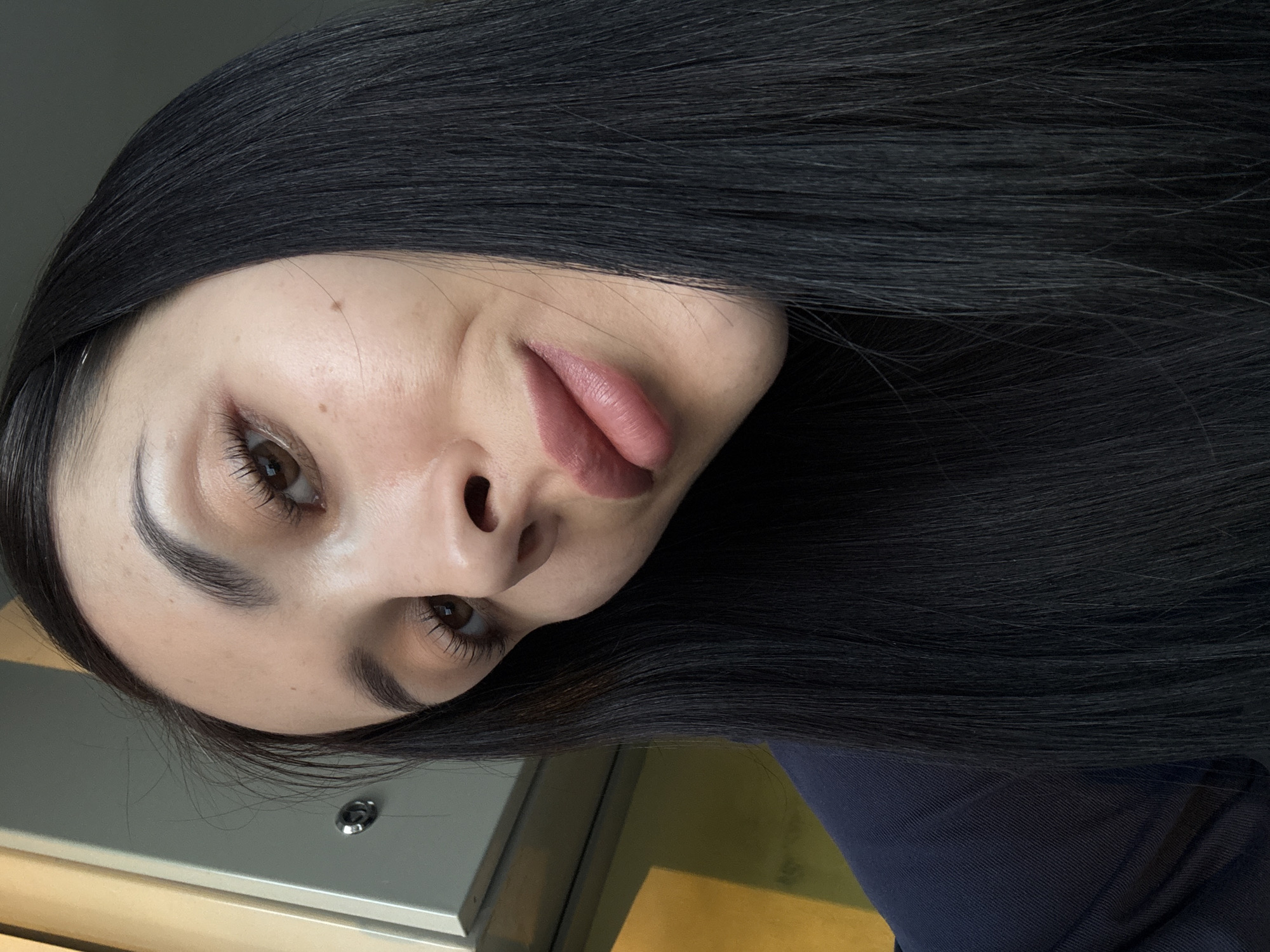 A close-up selfie of a fair-skinned young woman with long, dark hair. She has brown eyes, thick eyebrows, and is wearing lip color. The background is out of focus, but shows a neutral-colored surface with a metal button or knob, as well as a dark blue fabric. There is no indication of the location being in Molodezhnyi, Russia.