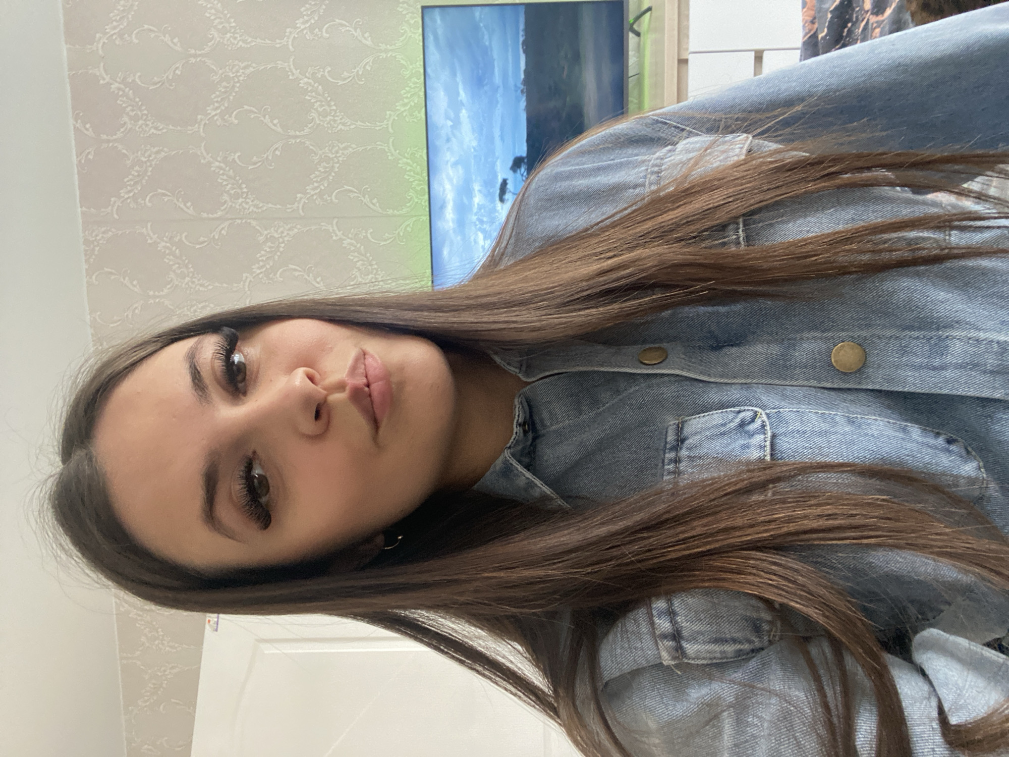 The image shows a woman with long, dark brown hair, wearing a blue denim jacket. She is looking at the camera with a slight pout. In the background, there is a patterned wallpaper and a television screen displaying a landscape scene. A white door is visible at the bottom of the frame. The location is Chaikovsky, Russia.