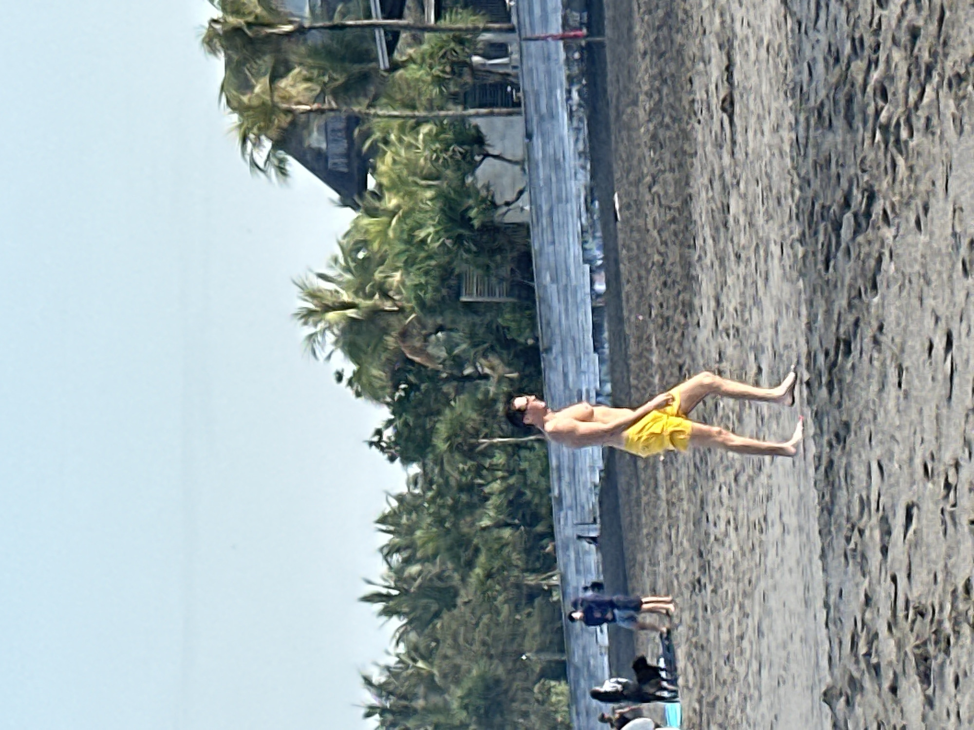 The image depicts a sunny day at a beach in North Kuta, Indonesia, with a focus on a man walking on the dark sand. The man is wearing bright yellow swim trunks and is shown from the knees up. He has short, dark hair and is wearing glasses. In the background, other people are visible, including a couple with a black dog, and individuals standing near some buildings and palm trees. A stone retaining wall is present behind the people, and a dark building with a thatched roof, partially obscured by palm trees, is visible further in the distance. The sky is clear and blue. The foreground is dominated by the textured, dark sand of the beach.