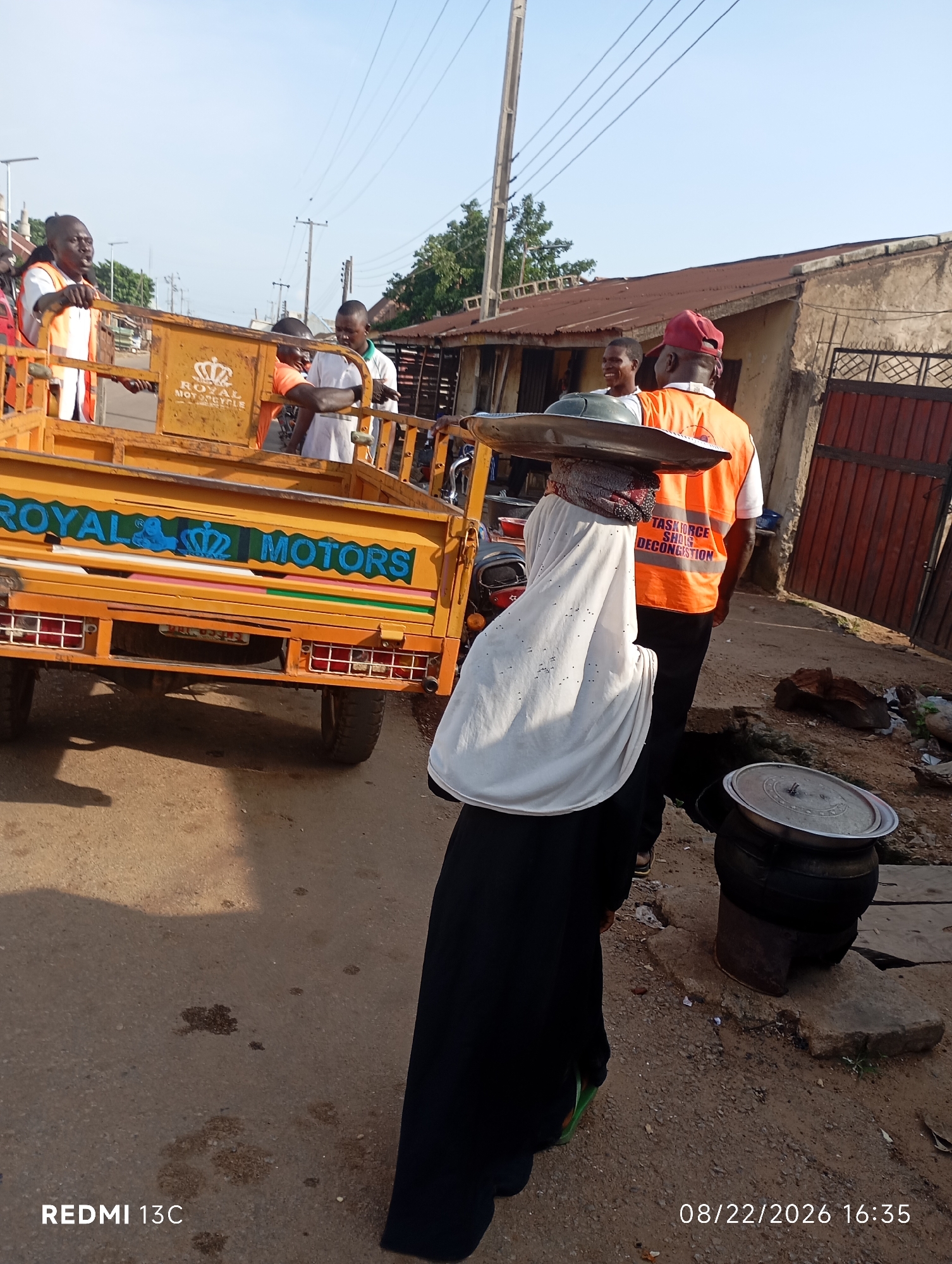 The image shows a street scene with several people and a yellow tricycle. In the foreground, a person wearing a white veil and a black abaya is walking away from the camera, carrying a large metal tray on their head. Behind this person, a man in an orange vest with "TASK FORCE SHAG DECONGESTION" written on the back is standing. The yellow tricycle has "ROYAL MOTORS" written on its side in blue letters. Several other people are visible in the background, some of whom appear to be near the tricycle, possibly engaged in a transaction or conversation. The environment appears to be a dusty street with buildings and some vegetation. The overall impression is a busy street with people involved in transportation and possibly commerce.