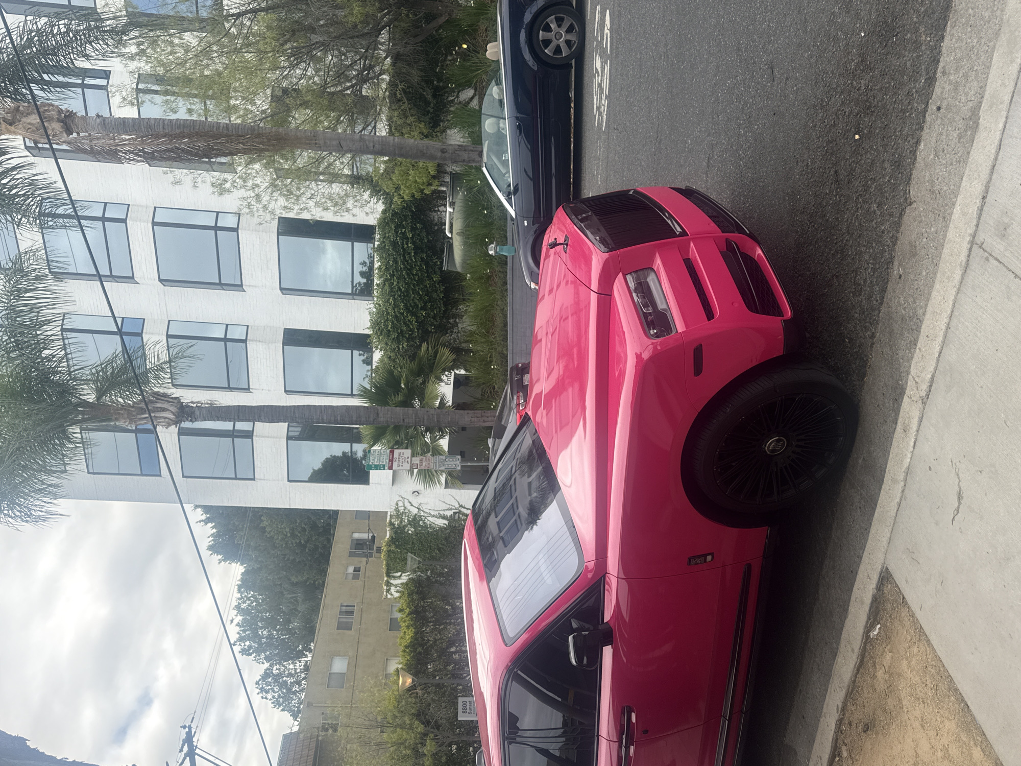 The image is taken at street level and shows a pink Hummer EV parked on a street in West Hollywood, USA. The vehicle is facing to the right of the frame and is parked next to a dark-colored sedan. In the background, there is a tall white building with large windows and a yellow apartment building. Palm trees line the street and the sky is overcast. There is a street sign visible near the white building.